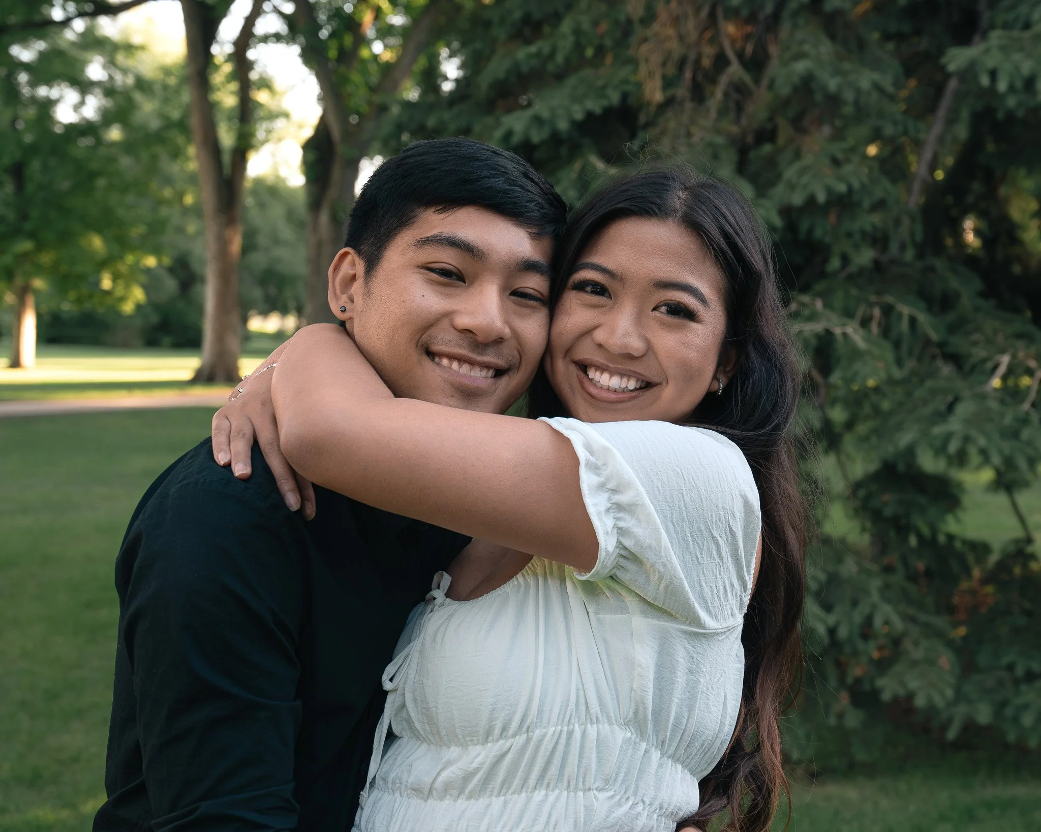 A young man and woman smiling and hugging outdoors in a park with trees in the background.