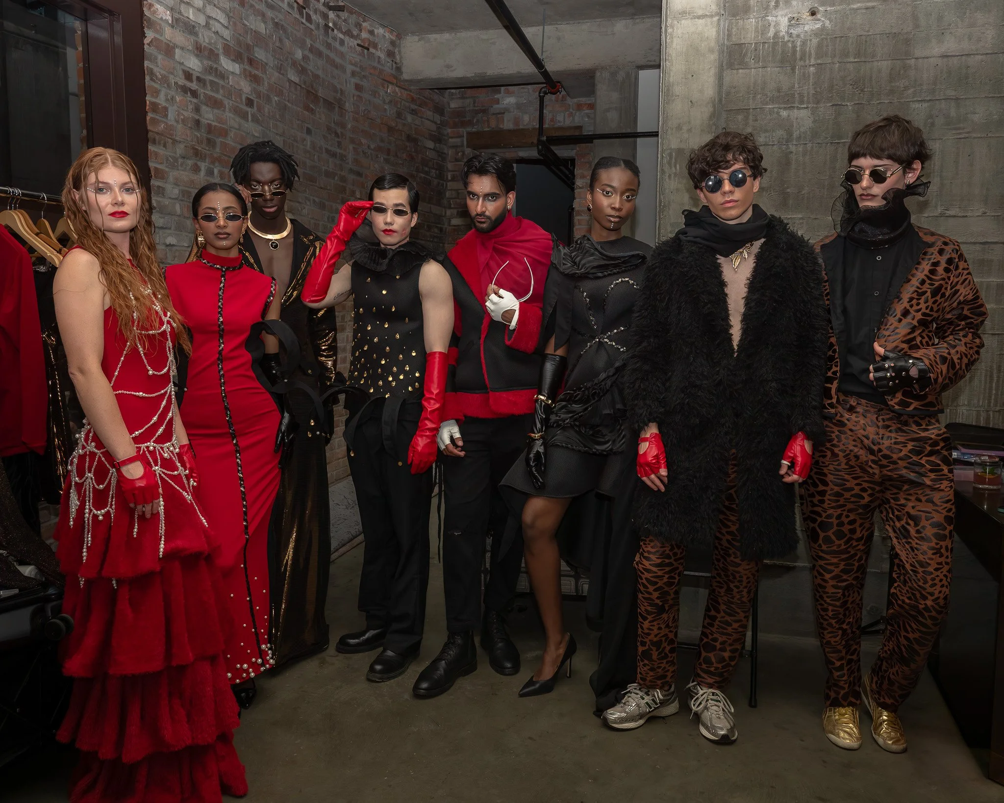 Group of people dressed in fashionable, bold, and eclectic outfits, standing together indoors against a brick and concrete wall, with some wearing sunglasses and dramatic makeup.