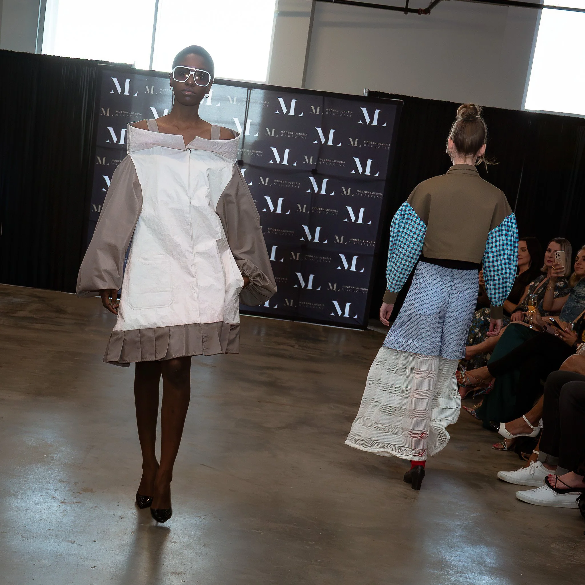 A fashion runway model walking in a beige and white layered outfit with large white sunglasses, while an audience watches and takes photos in the background.