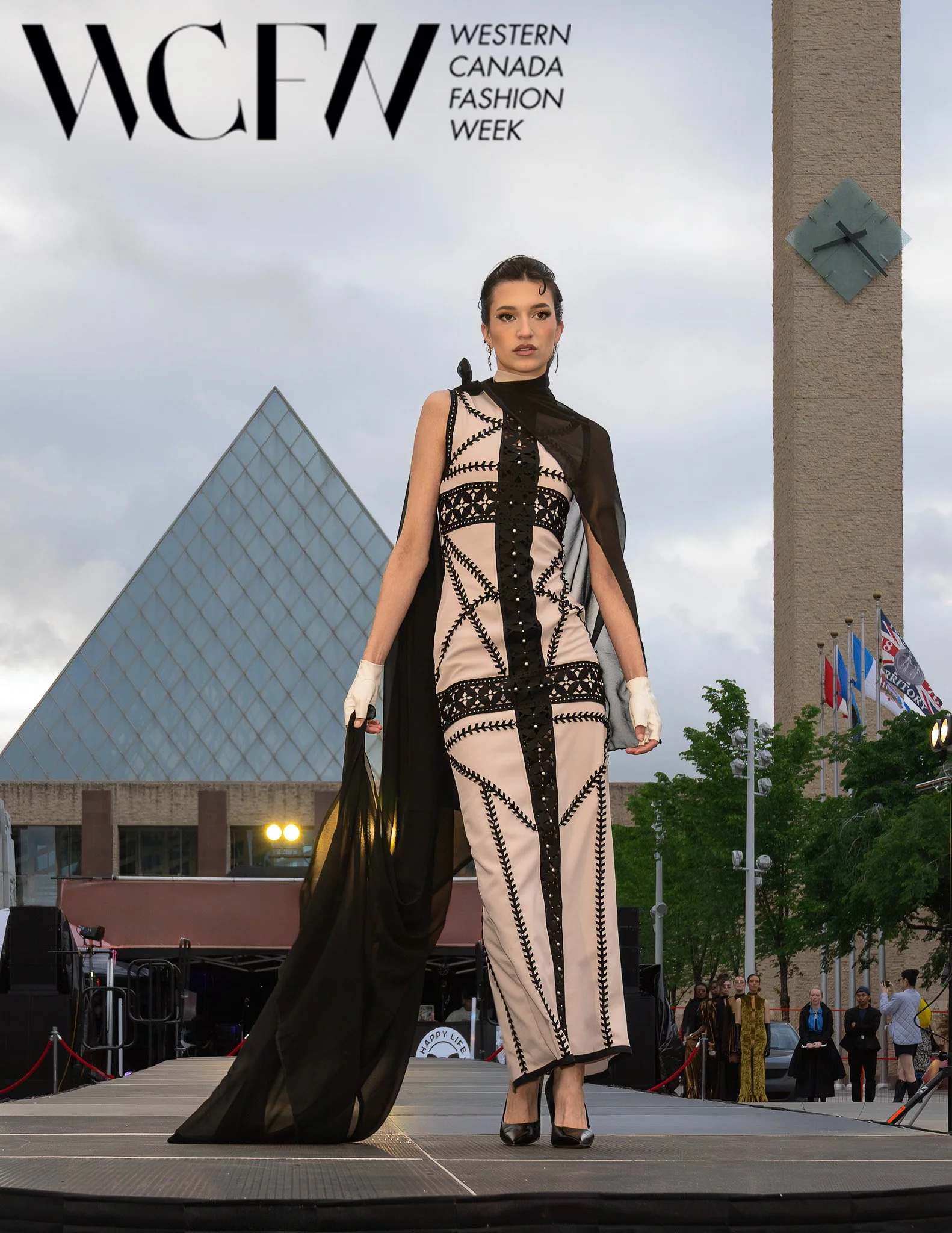 Model walking on runway at an outdoor fashion show during Western Canada Fashion Week, with audience and flags in the background, and a modern building with a pyramid-shaped glass roof behind.