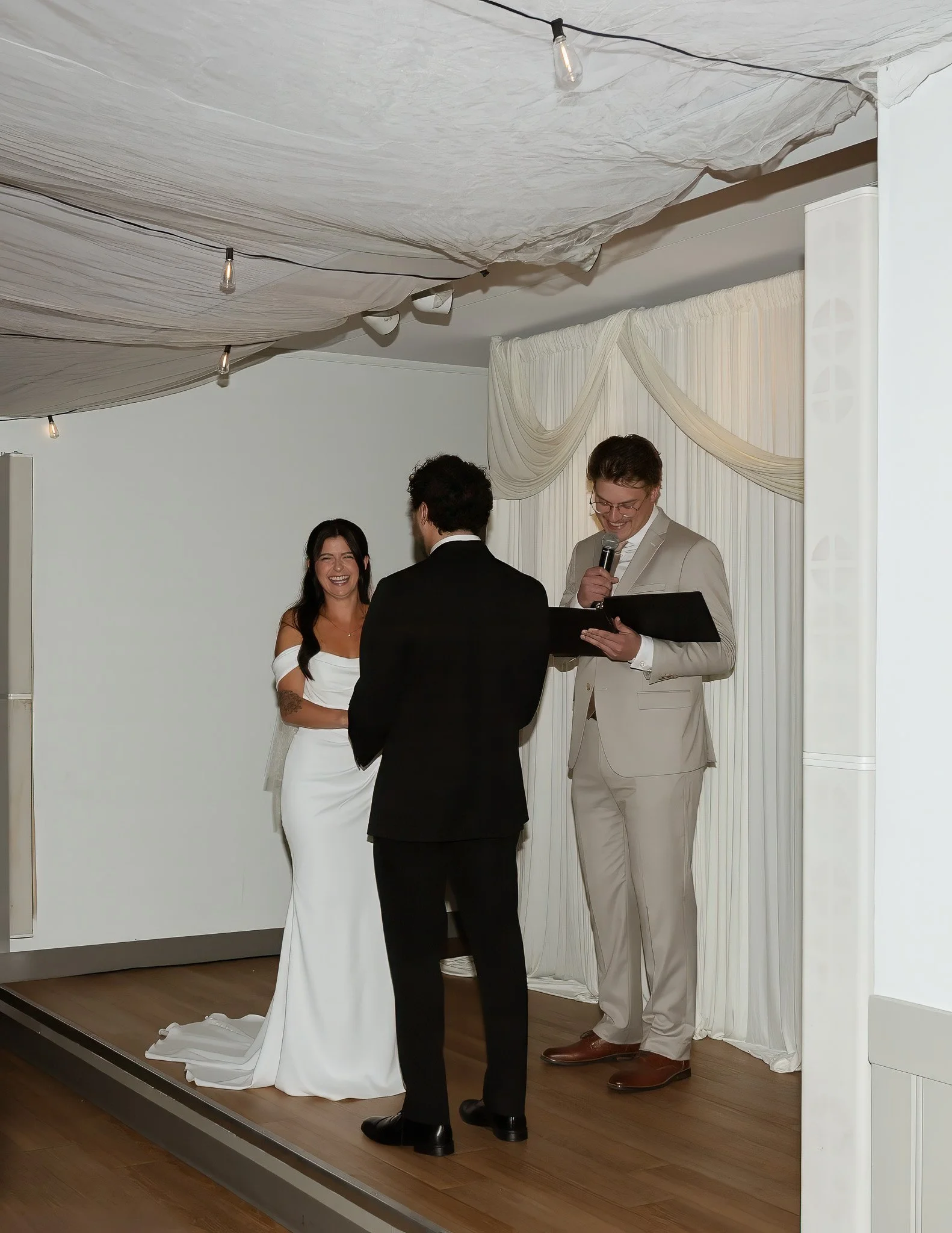 A wedding ceremony with four people on a small stage, including a bride in a white dress, a groom in a black suit, an officiant in a beige suit, and another person in a black suit. The ceremony is taking place indoors with white draped curtains and s