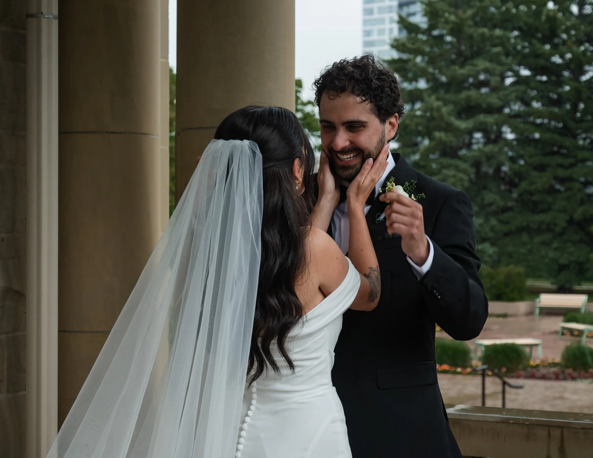 A bride and groom smiling closely outdoors during ceremony, bride wearing a white dress with veil, groom in a black tuxedo with boutonniere, touching faces, surrounded by trees and architectural columns.