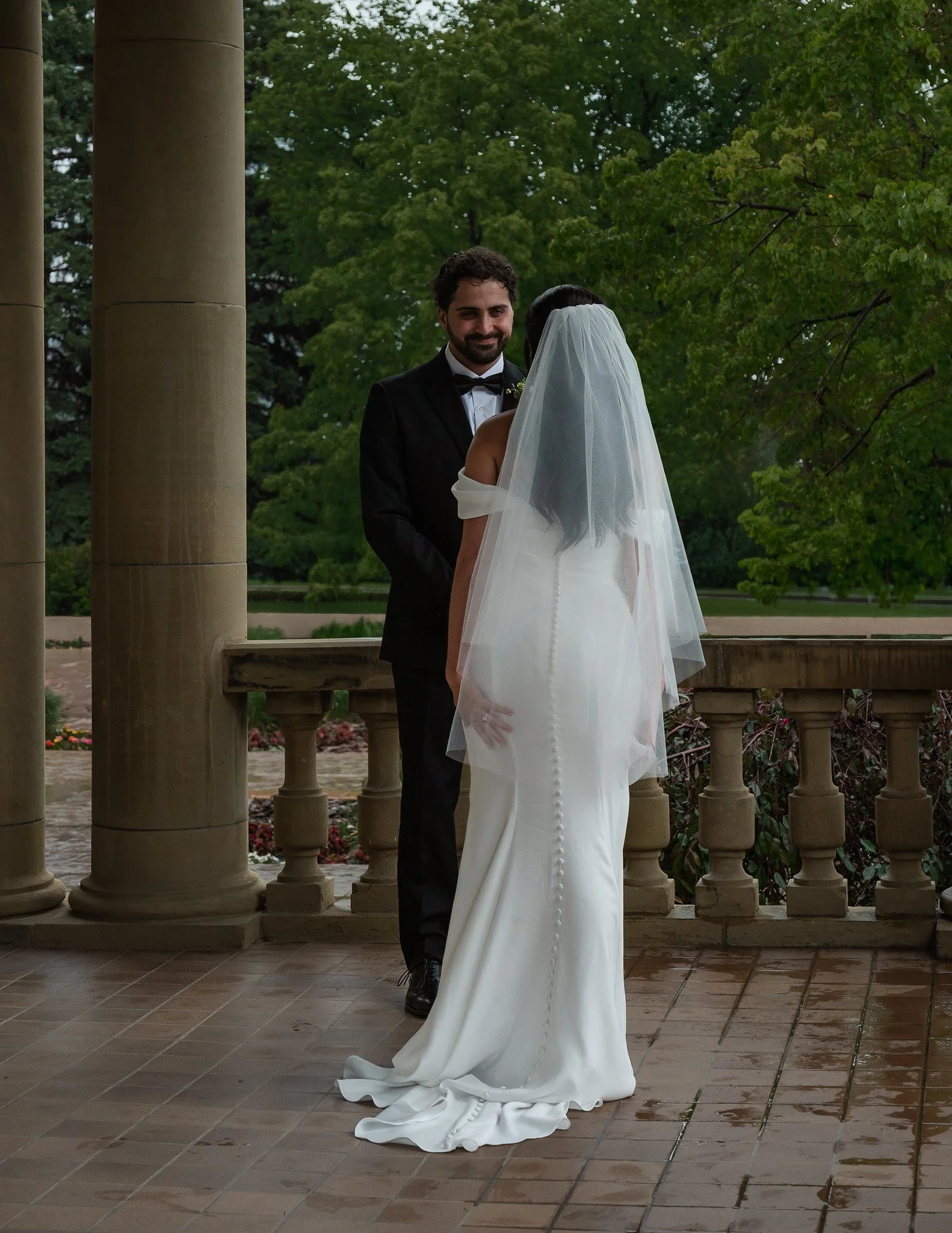A bride and groom standing under a pavilion with green trees in the background. The groom is smiling and wearing a black tuxedo with a bowtie, and the bride is wearing a white wedding gown with a veil. The scene is outdoors, and the ground appears we