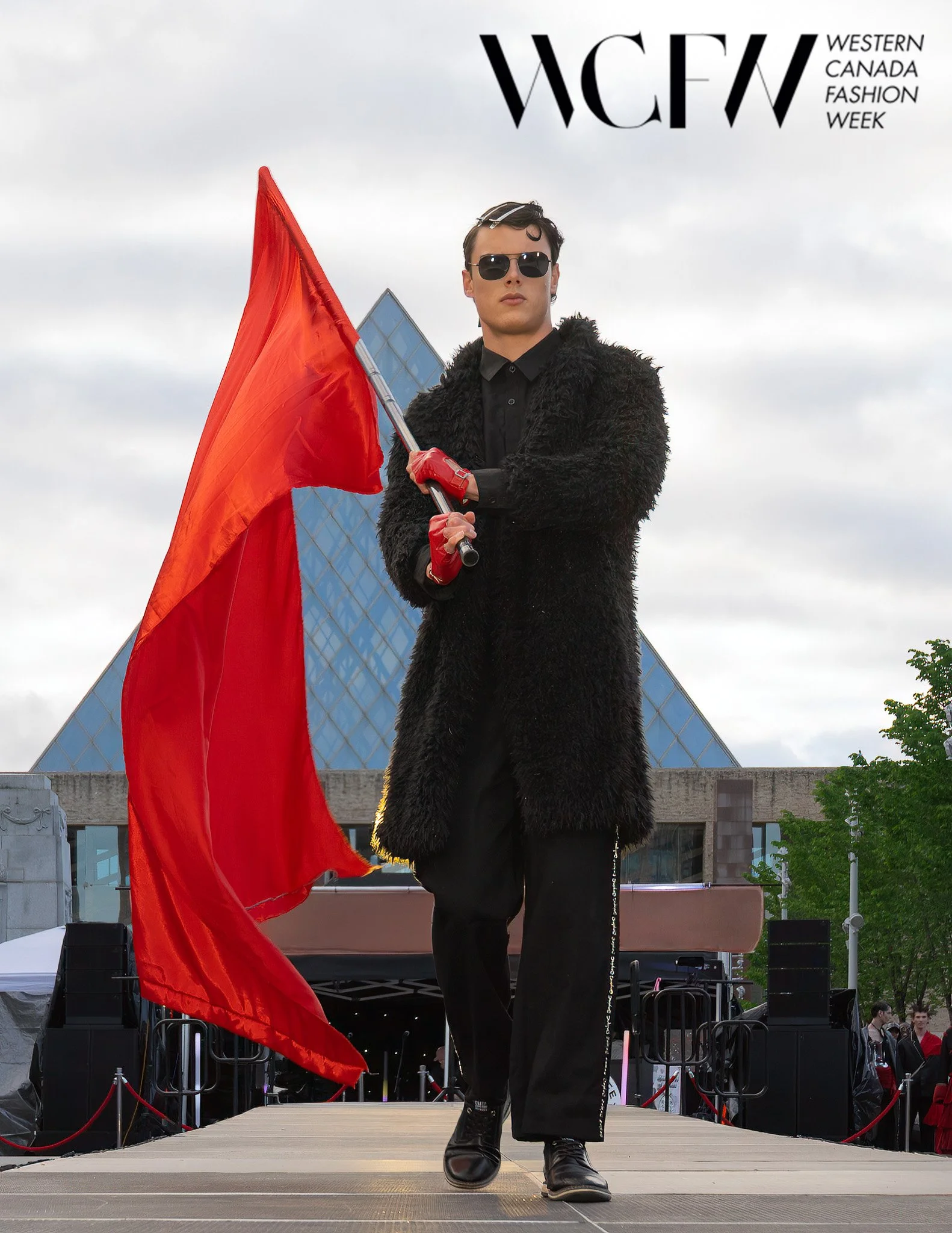 Young man walking down runway during Western Canada Fashion Week, holding a large red flag, wearing sunglasses, a black fur coat, black pants with white text, and black boots, with a glass building and cloudy sky in the background.