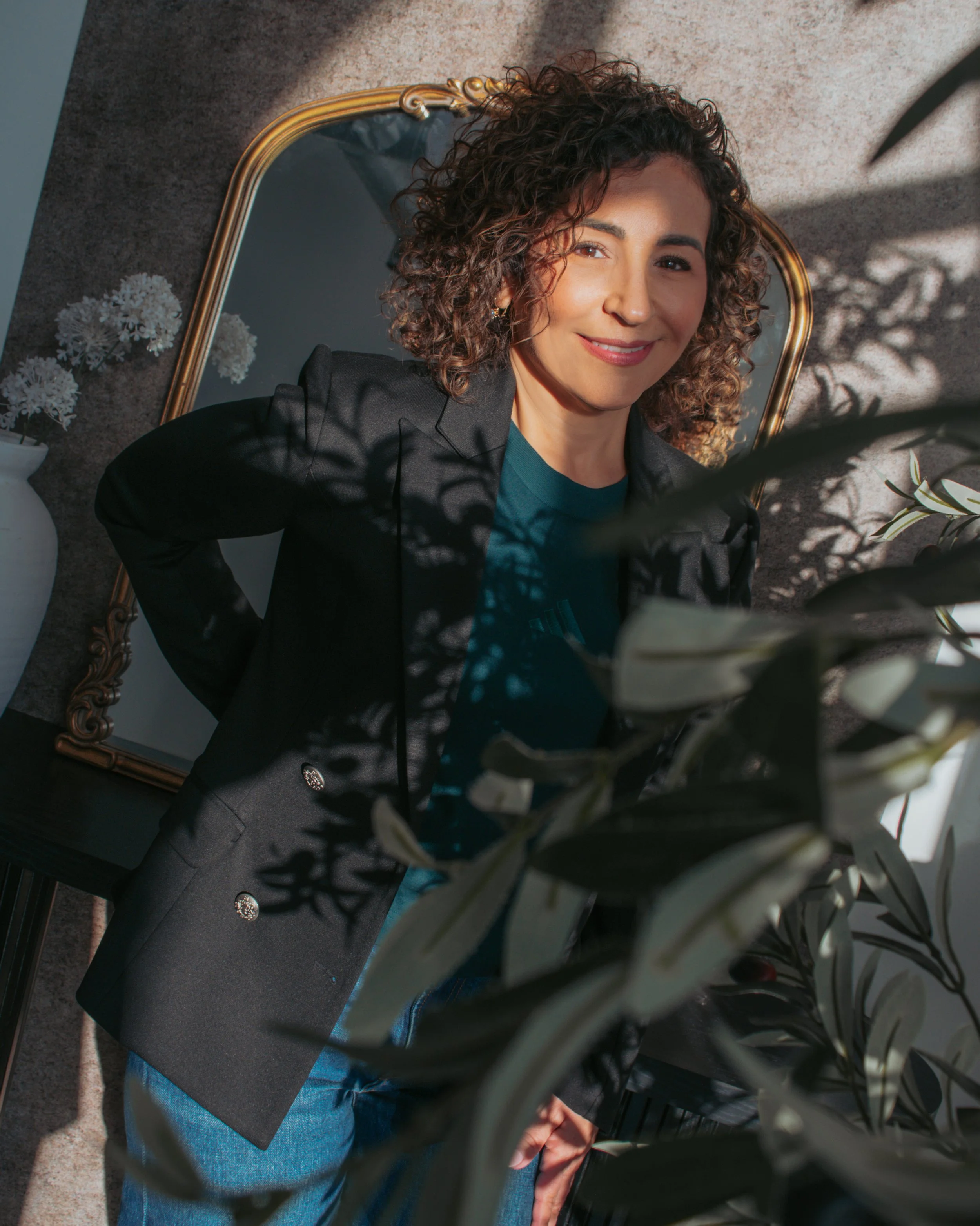 Woman with curly hair smiling and looking at camera, standing in front of a mirror, surrounded by plants and flowers in a room with natural light.