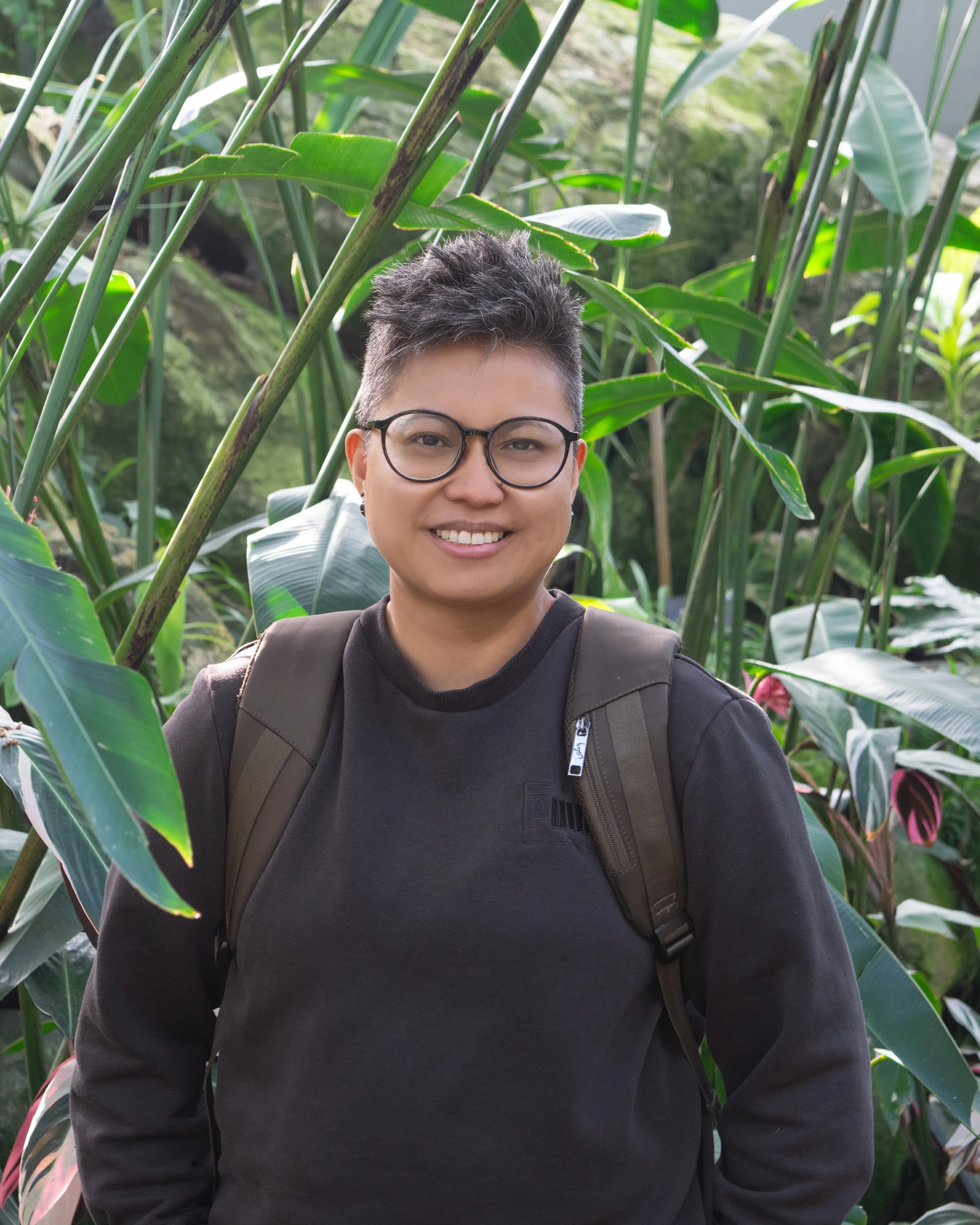 Person with short hair, glasses, and a black sweatshirt standing among green plants in a lush outdoor setting, smiling at the camera.