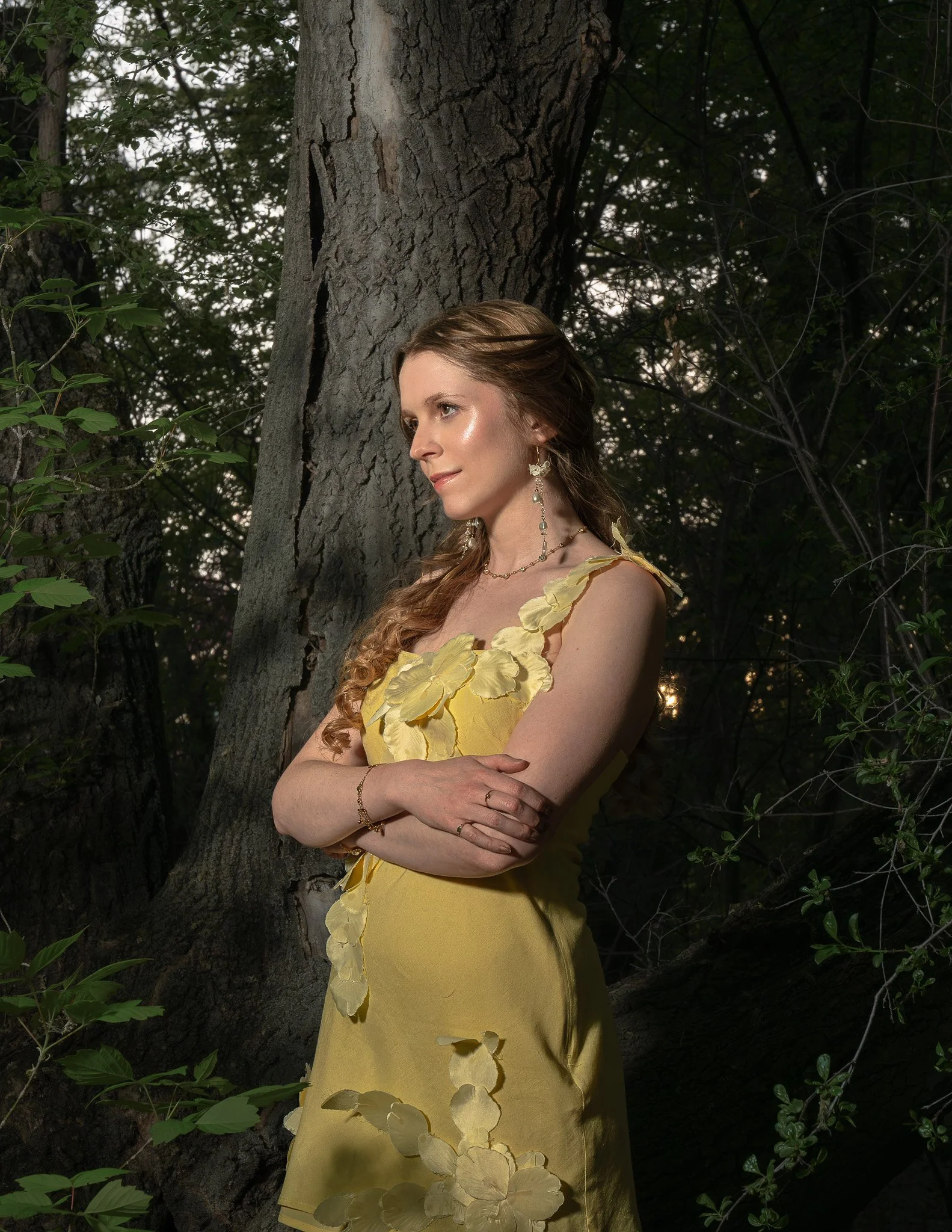 A woman with long, wavy, brown hair standing outdoors in a forest near a tree. She is wearing a yellow dress with petal-like details and jewelry, and she is looking to the side with arms crossed.
