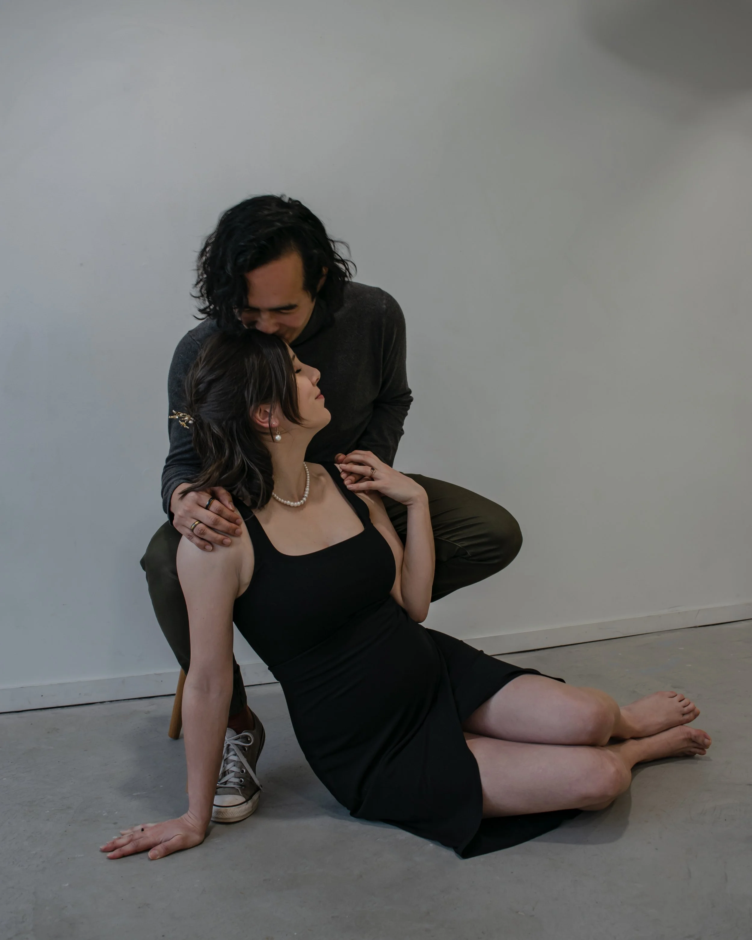A man with shoulder-length dark hair is kneeling behind a woman seated on the floor. The woman is wearing a black dress, pearl necklace, and earrings, with her dark hair styled in waves. She appears to be looking up at the man. The man is wearing a d
