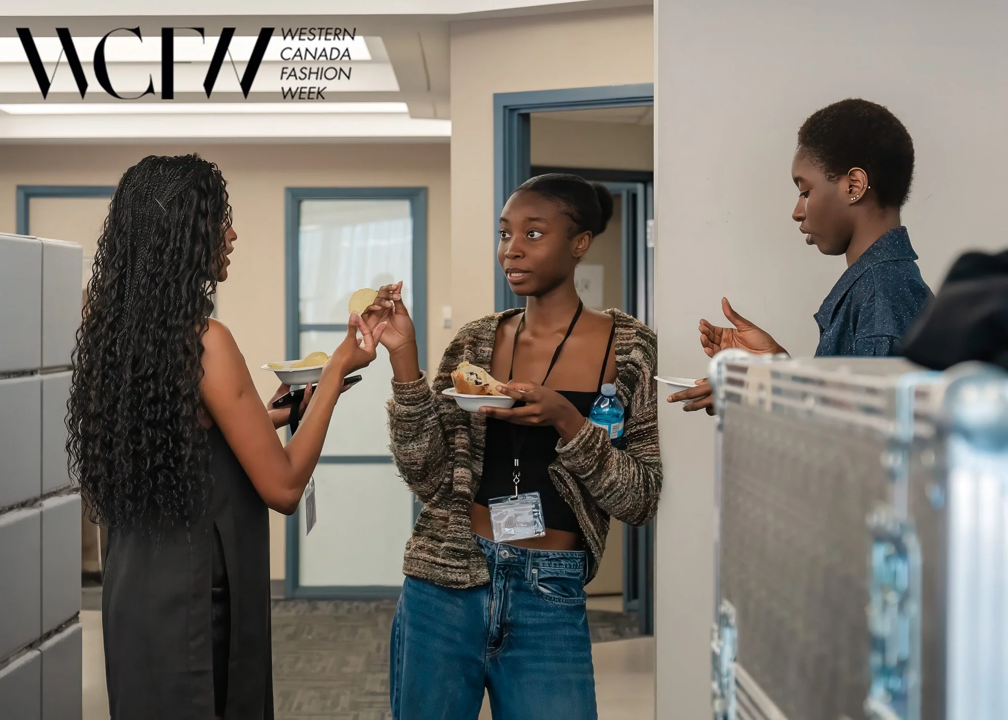 Three women having a conversation in an office, with a sign that reads 'Western Canada Fashion Week' in the background.