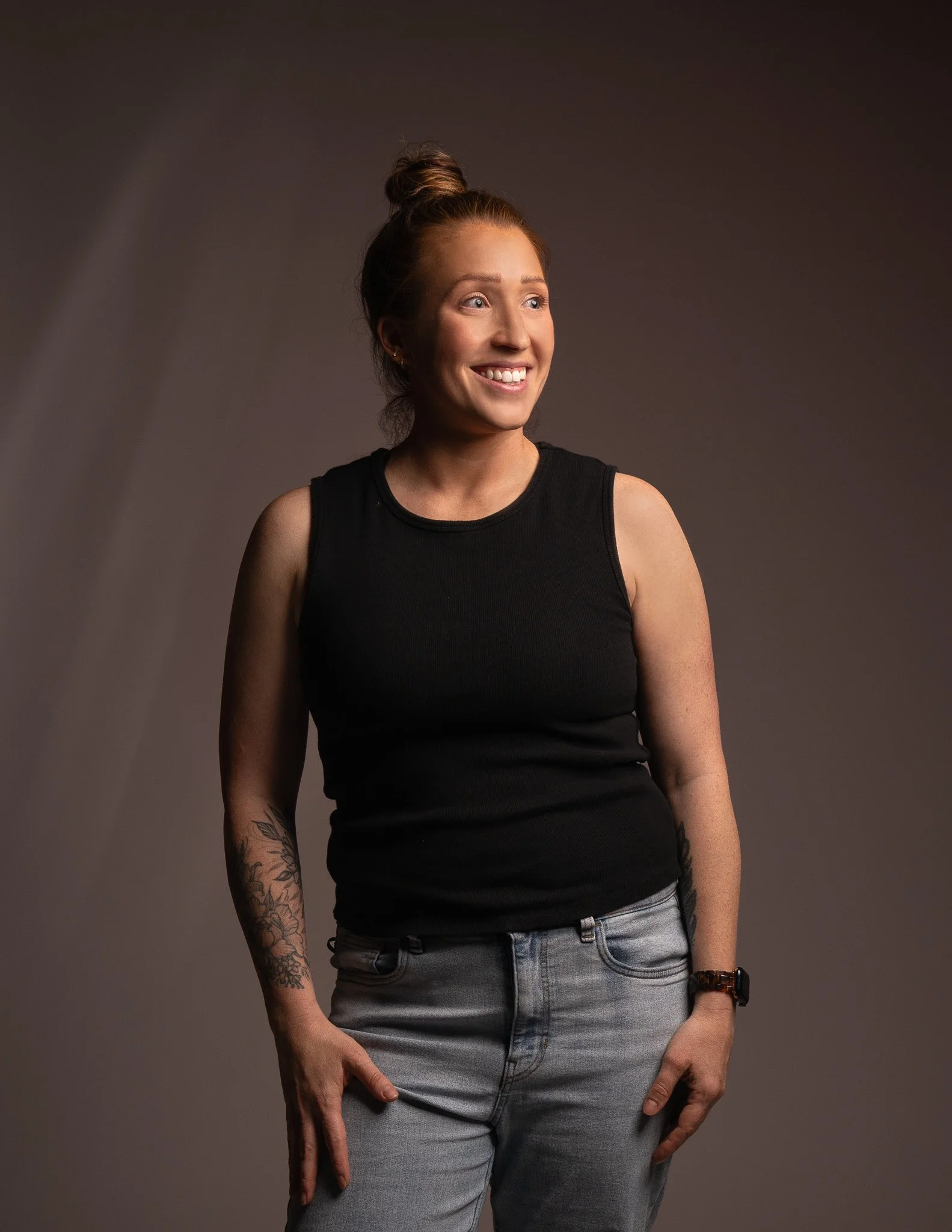 A woman with red hair tied in a bun, wearing a black sleeveless top and grey jeans, smiling and looking to her right, with tattoos on her left arm, against a dark neutral background.