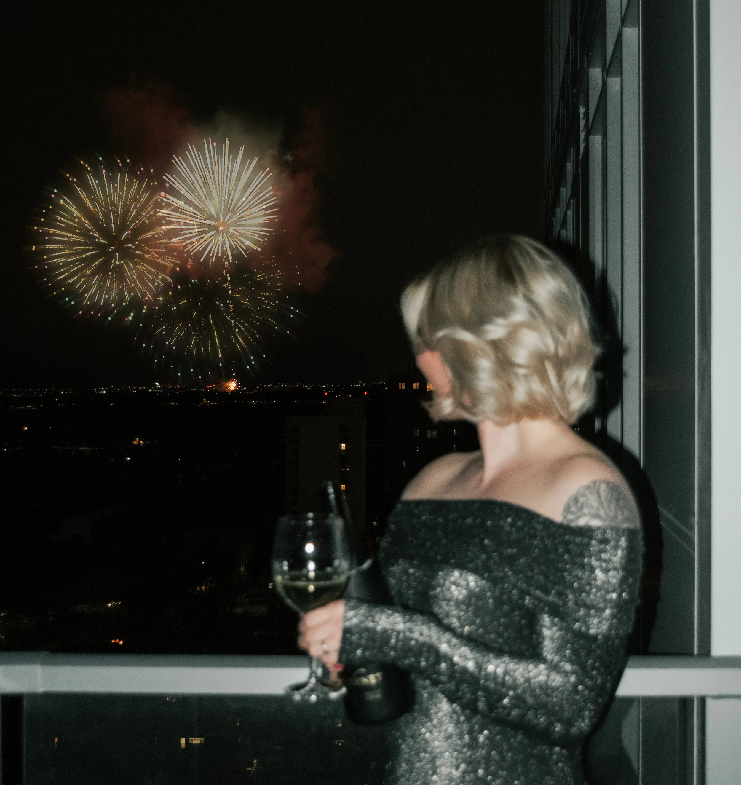 Woman with short blonde hair and tattoos, wearing a sparkly off-the-shoulder dress, holding a glass of white wine, watching fireworks display in the night sky from a balcony.