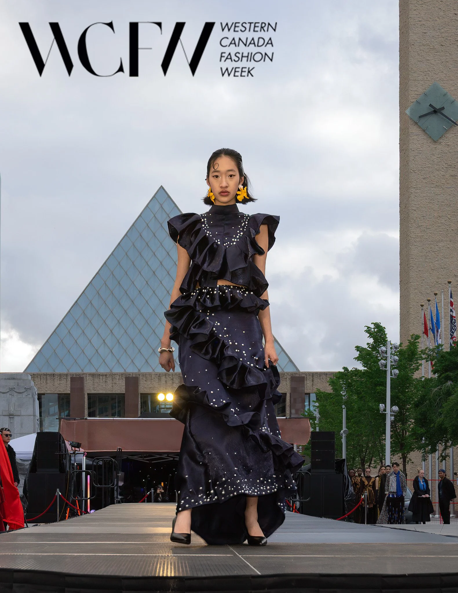 Model walking on runway during Western Canada Fashion Week, dressed in black ruffled gown with pearl embellishments, wearing large yellow flower earrings, with an audience and city buildings in background.