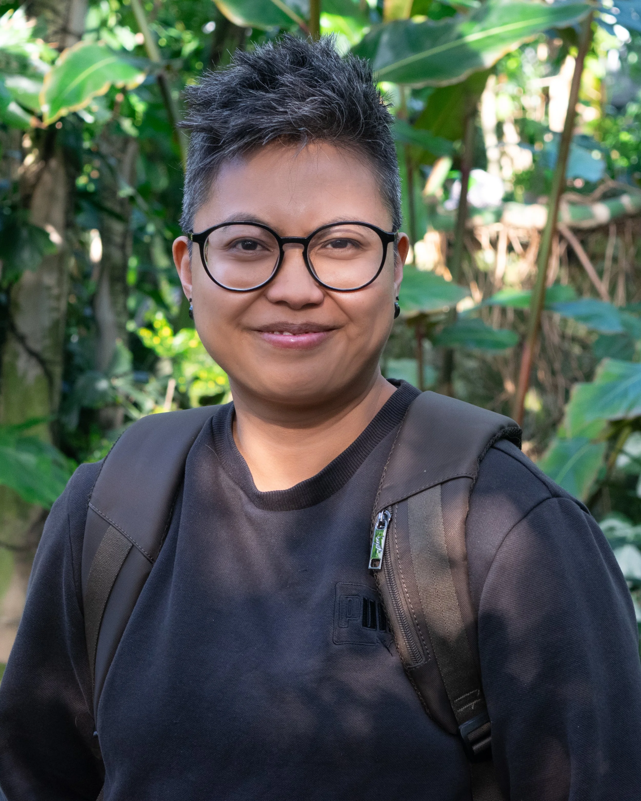 A person with short dark hair, glasses, and earrings, smiling outdoors with lush green foliage in the background.