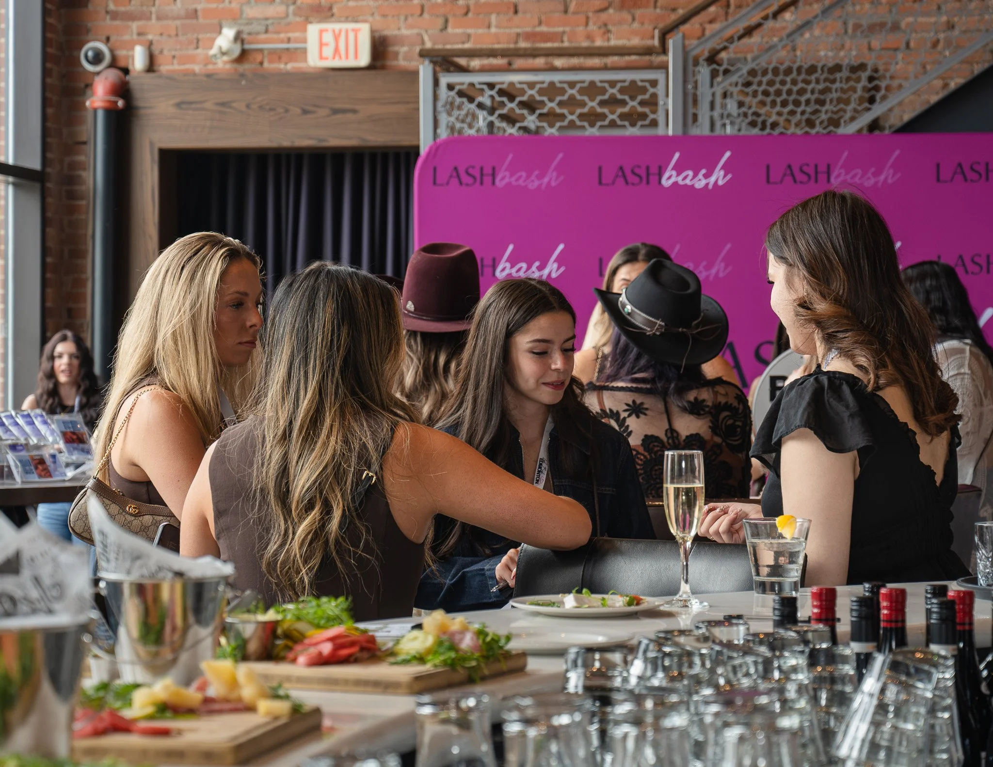 Group of women gathered around a table with food and drinks, at what appears to be a social event, with a pink backdrop reading 'LASH bash' and a brick wall in the background.