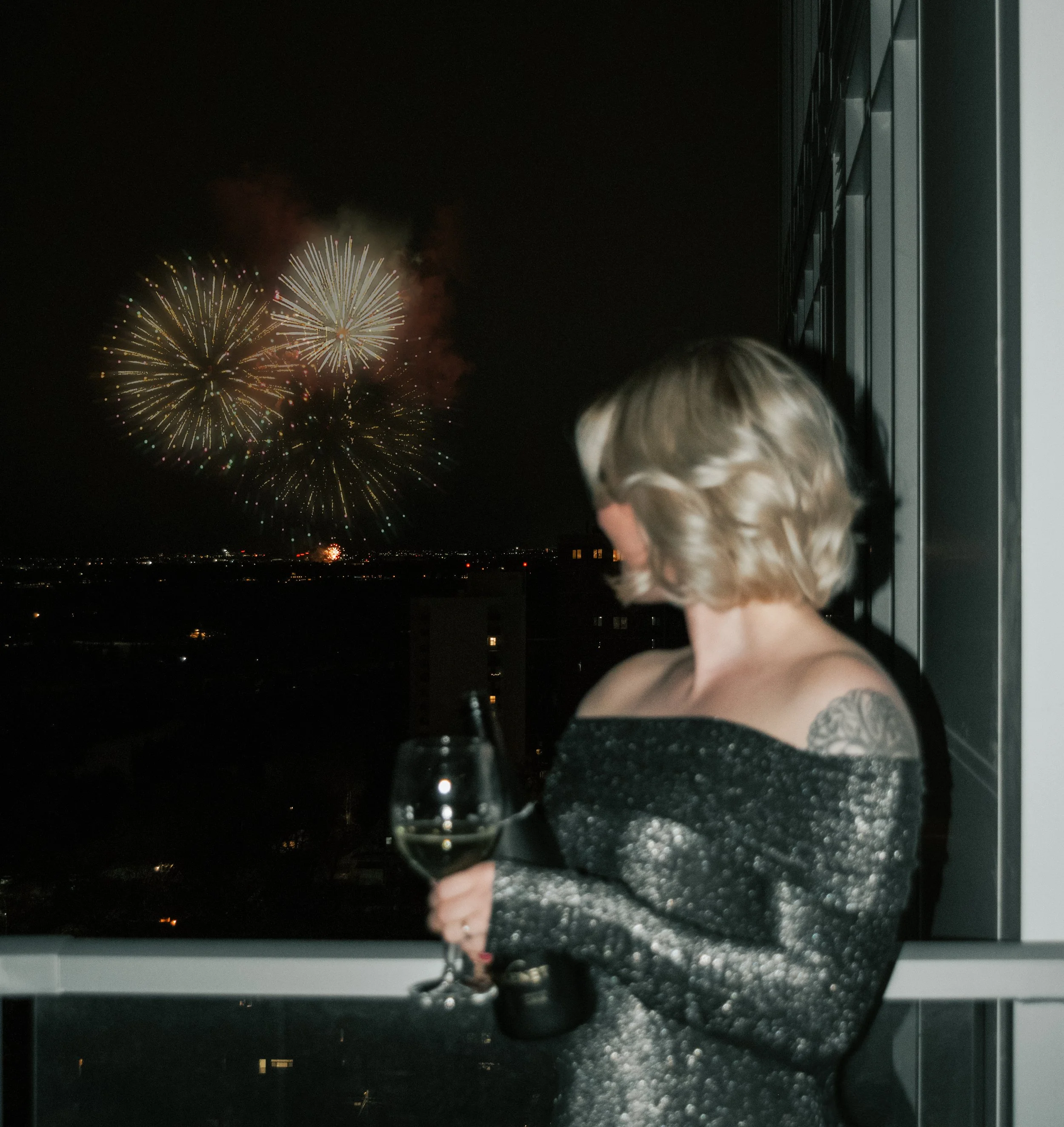 A woman with short blonde hair holding a glass of white wine, looking out at fireworks in the night sky from a balcony.