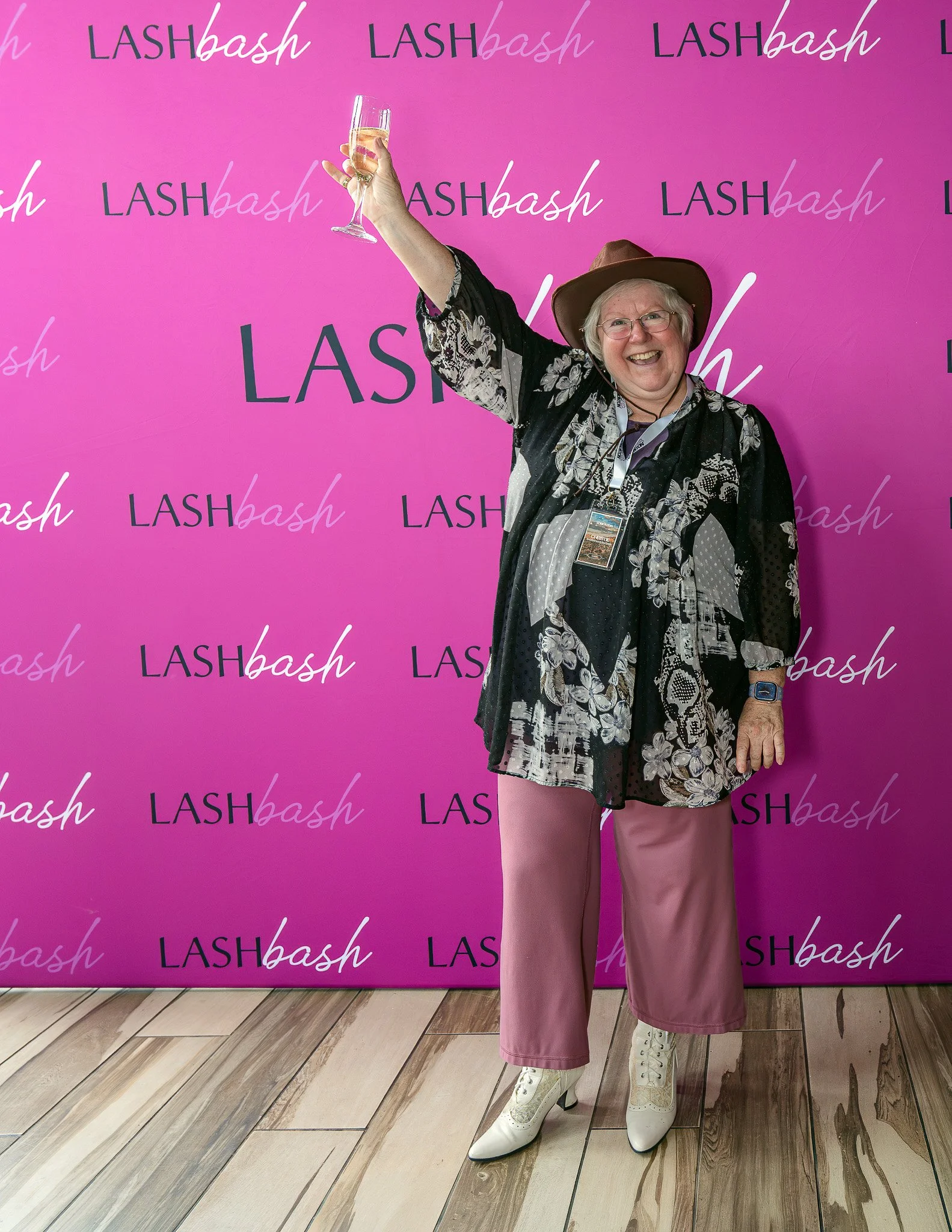 A smiling woman wearing glasses, a brown hat, a black and floral patterned cover-up, pink wide-leg pants, and white high-heeled cowboy boots, holding a glass of champagne in front of a pink backdrop with "LASHbash" written repeatedly in elegant scrip