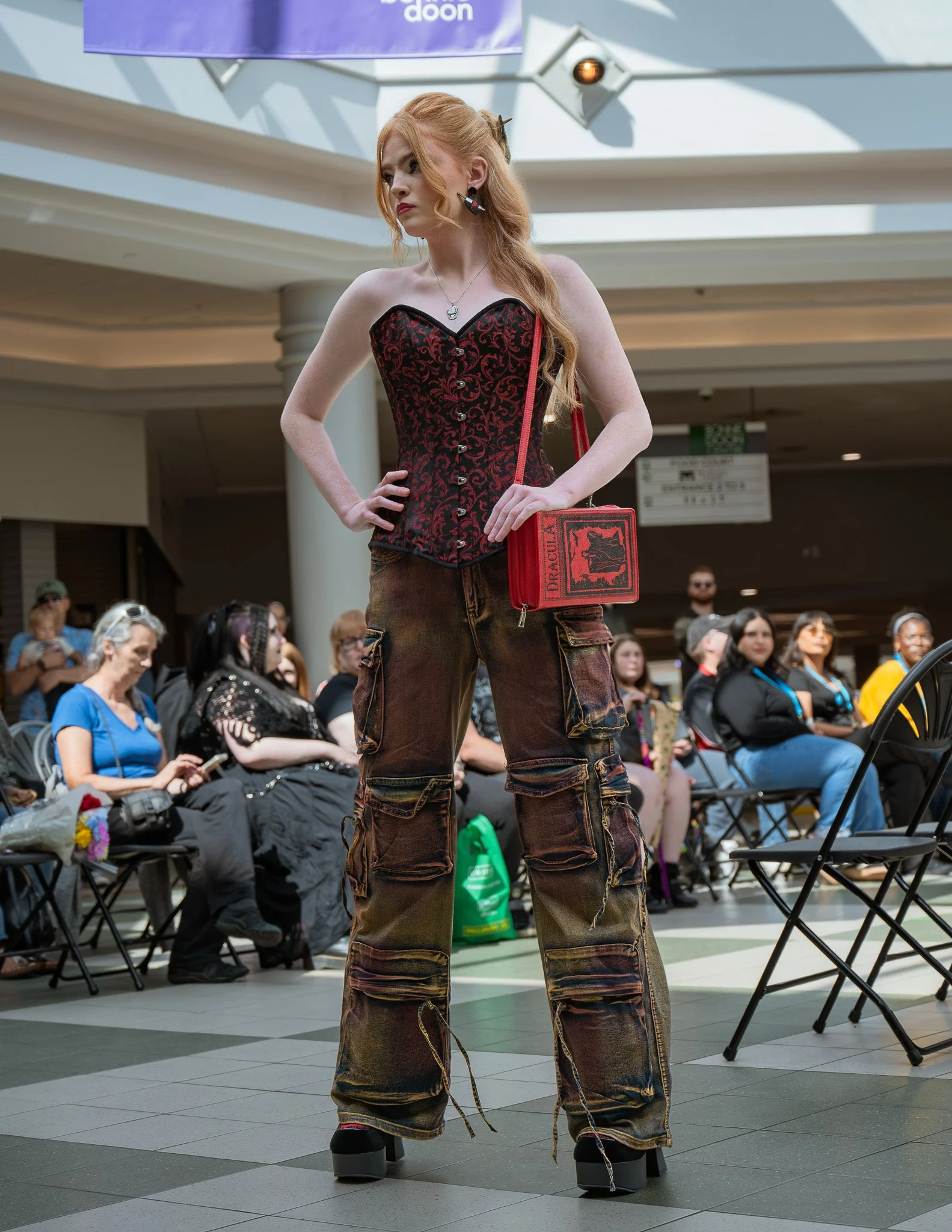 A woman with long red hair styled in loose waves, wearing a red and black patterned corset top, dark cargo pants, and platform shoes, standing with her hands on her hips in a shopping mall or public space. She has large earrings and a pendant necklace, and is carrying a red handbag with black graphic elements. There is a crowd of people sitting and standing in the background, and some are looking at her.