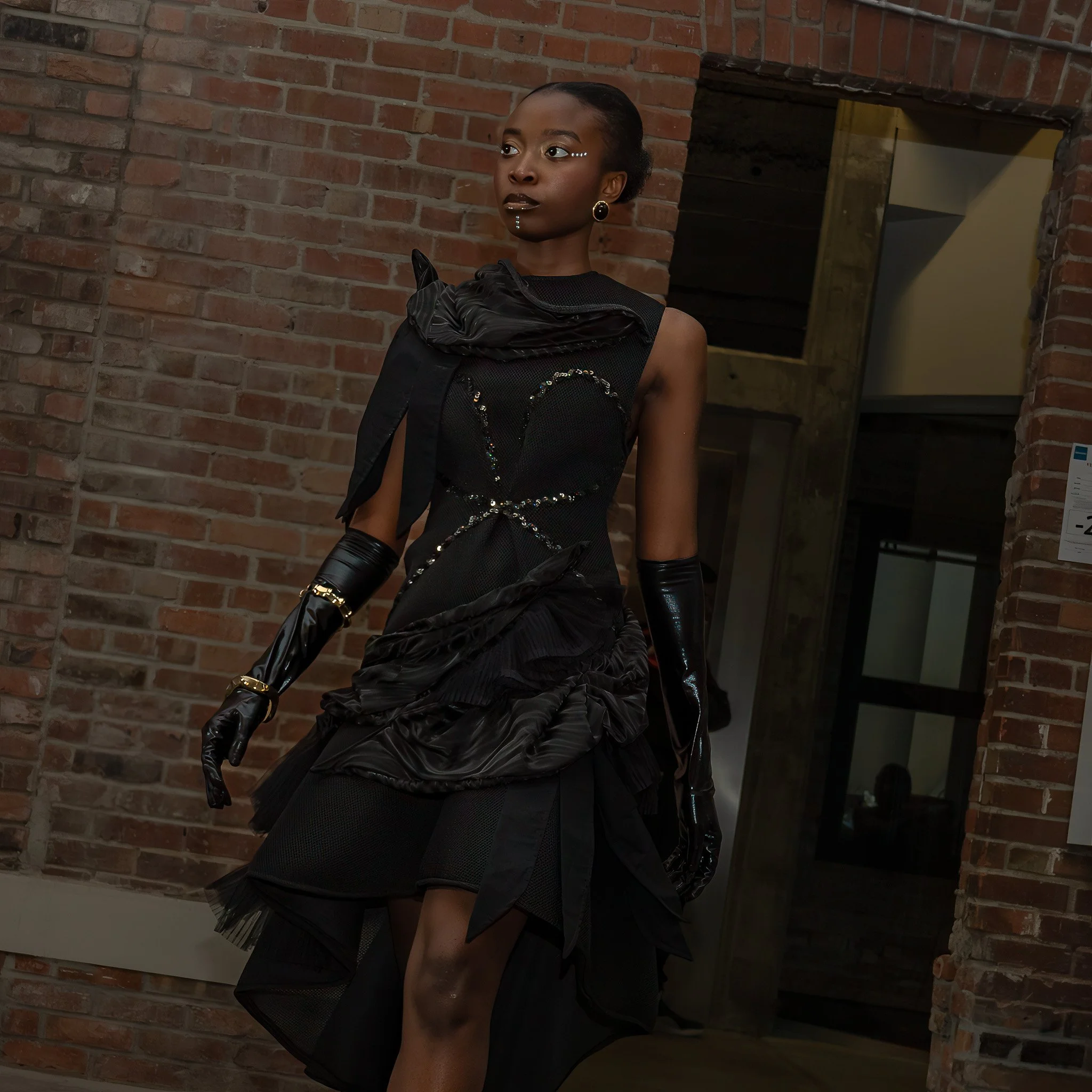 A woman wearing black, shiny gloves, and artistic makeup, standing against a brick wall with a mirror and some furniture in the background.