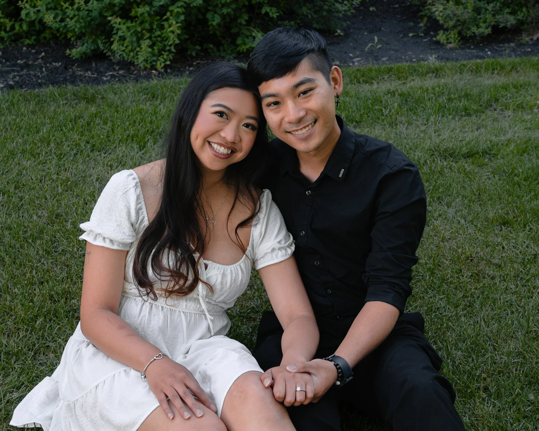 A smiling woman and man sitting closely together on the grass outdoors, with green bushes behind them.