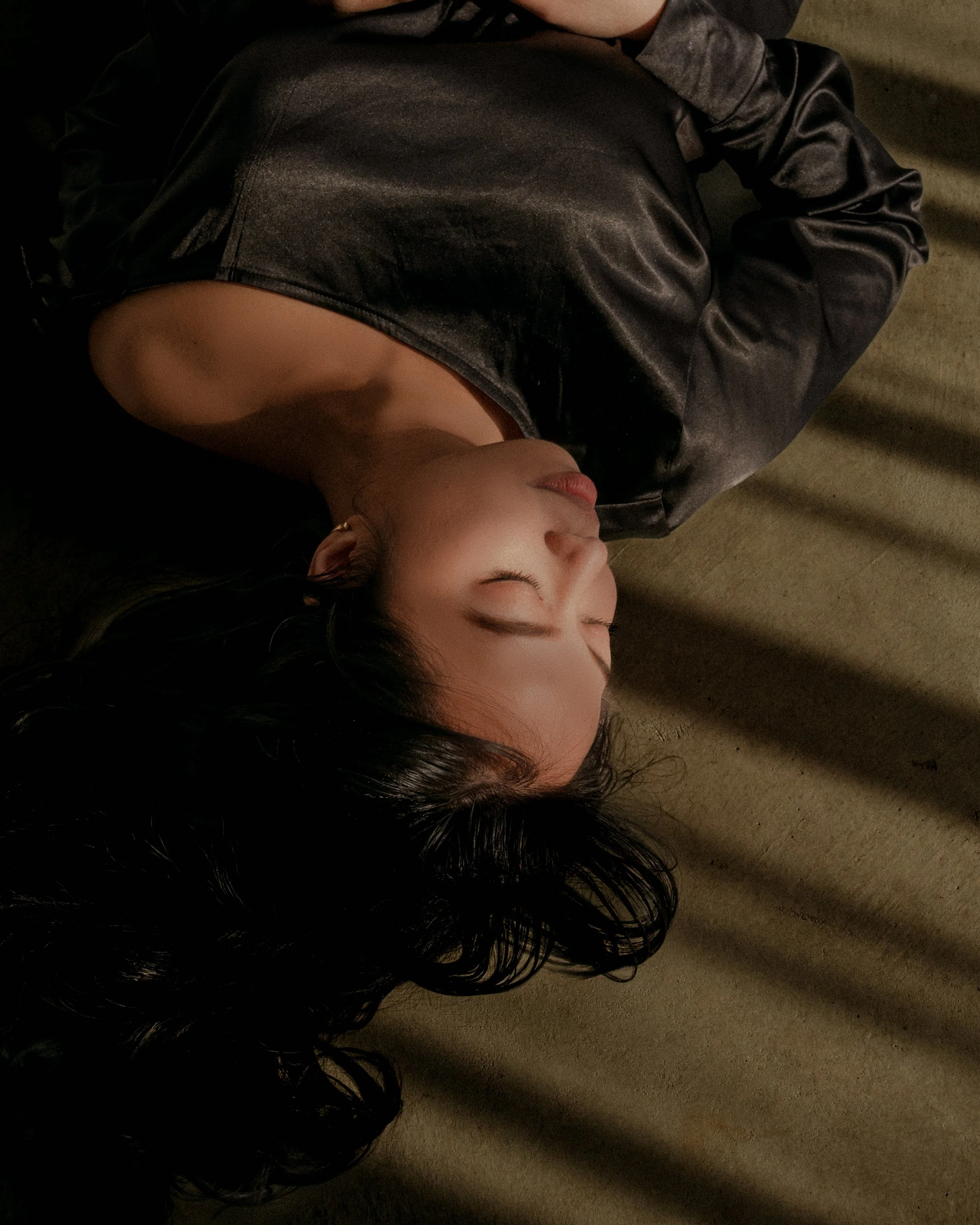 A woman lying on a beige carpeted floor with her eyes closed, wearing a shiny black satin top, and shadows cast across her face and body.