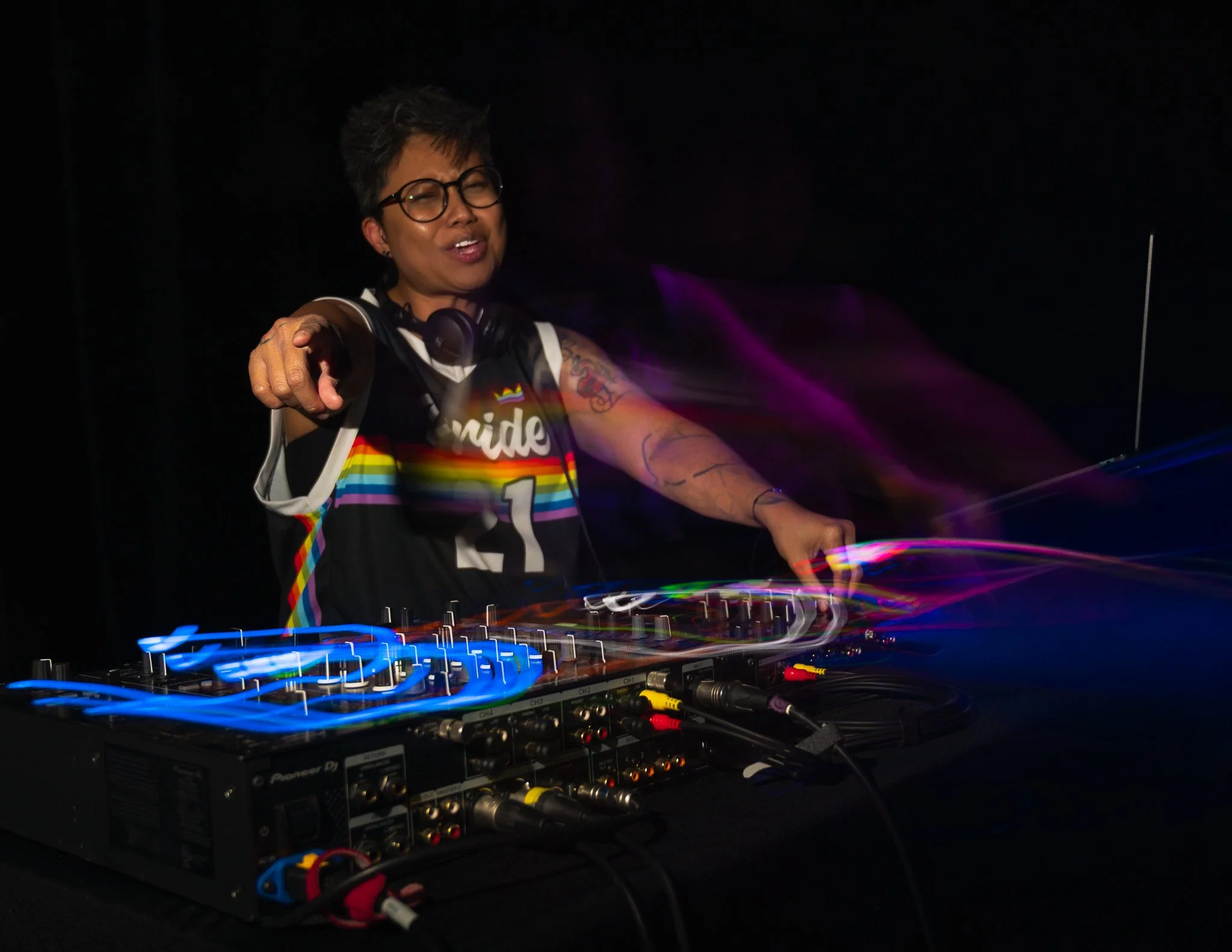 A DJ with short hair, glasses, and tattoos, wearing a rainbow-striped jersey, is performing with DJ equipment in a dark setting, illuminated by colorful lights.