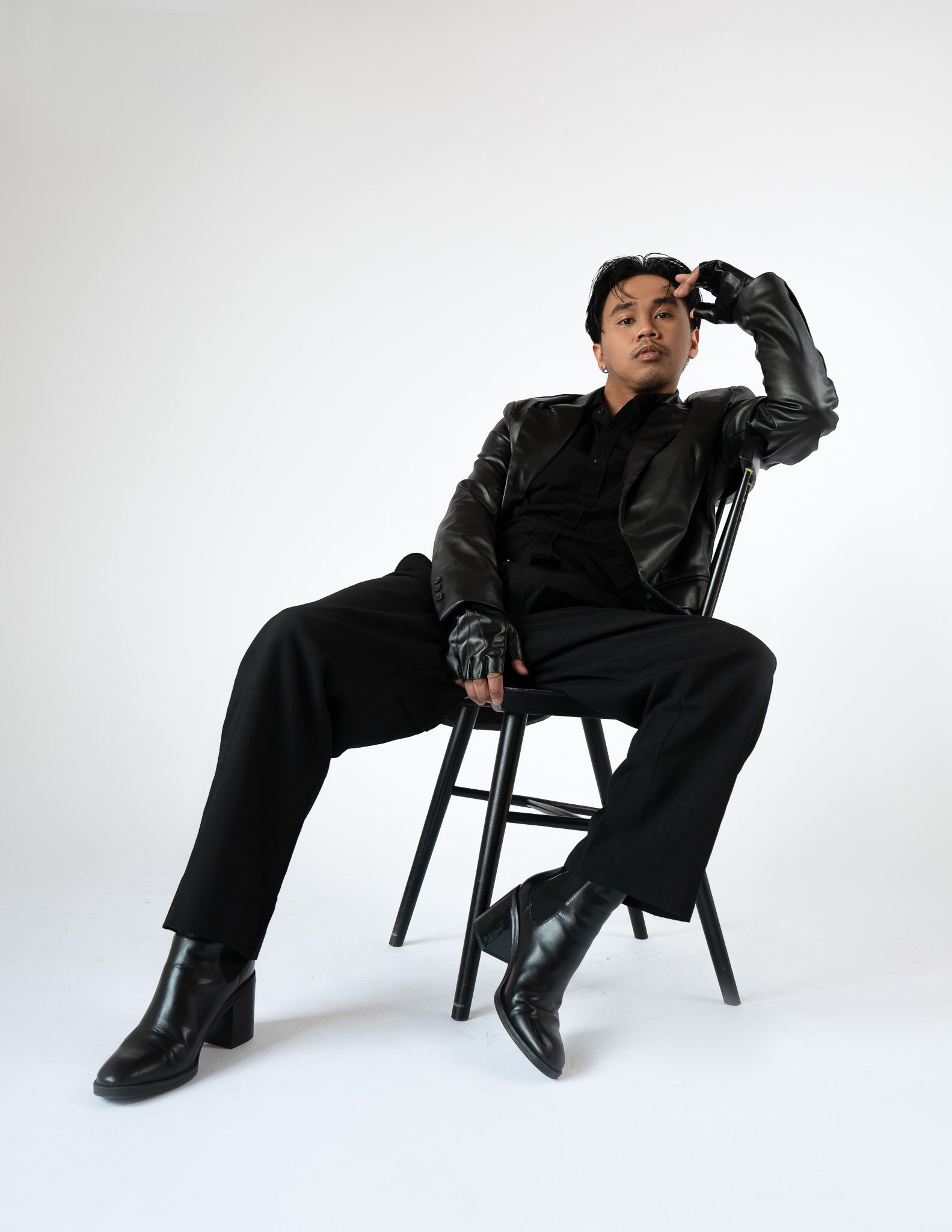 A person sitting on a black chair against a plain white background, dressed in black leather jacket, black shirt, black pants, and black high-heeled boots, with one hand resting on their head and the other on their thigh.
