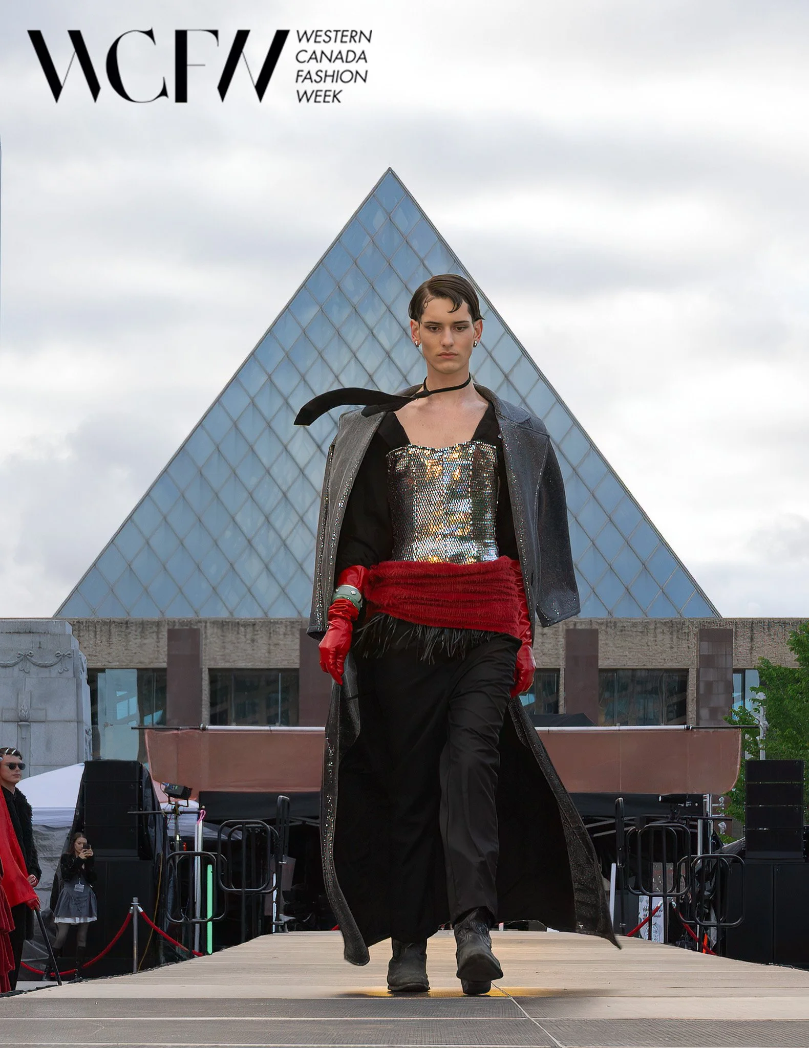 A fashion runway model walks on stage at Western Canada Fashion Week, with photographers and spectators on the sides. The backdrop features a large glass pyramid structure, and the model is dressed in a shiny top, long coat, red gloves, and black boo