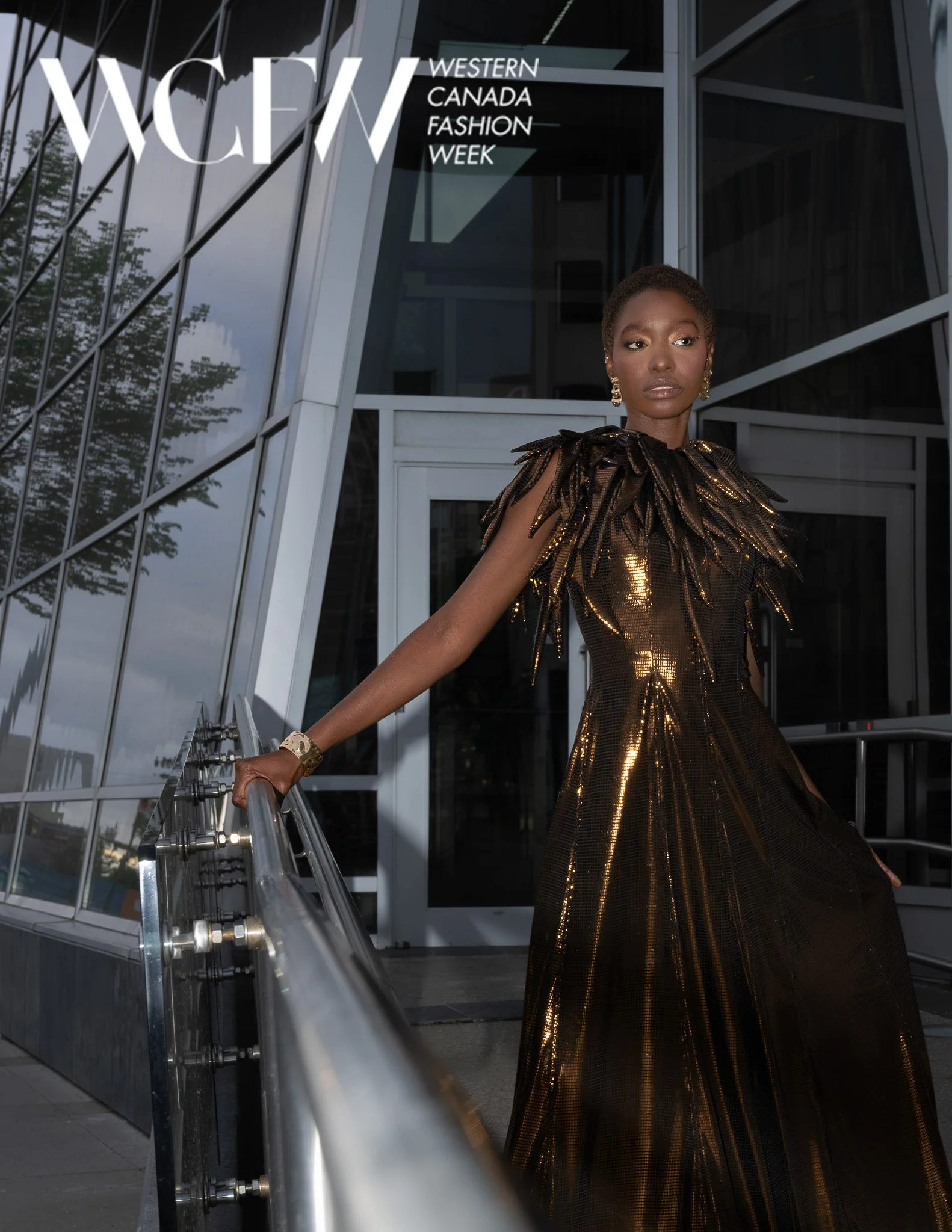 A woman wearing a shiny, metallic gold evening gown with black details and large earrings standing outside a modern building with a sign that says "Western Canada Fashion Week" and "WCFW."