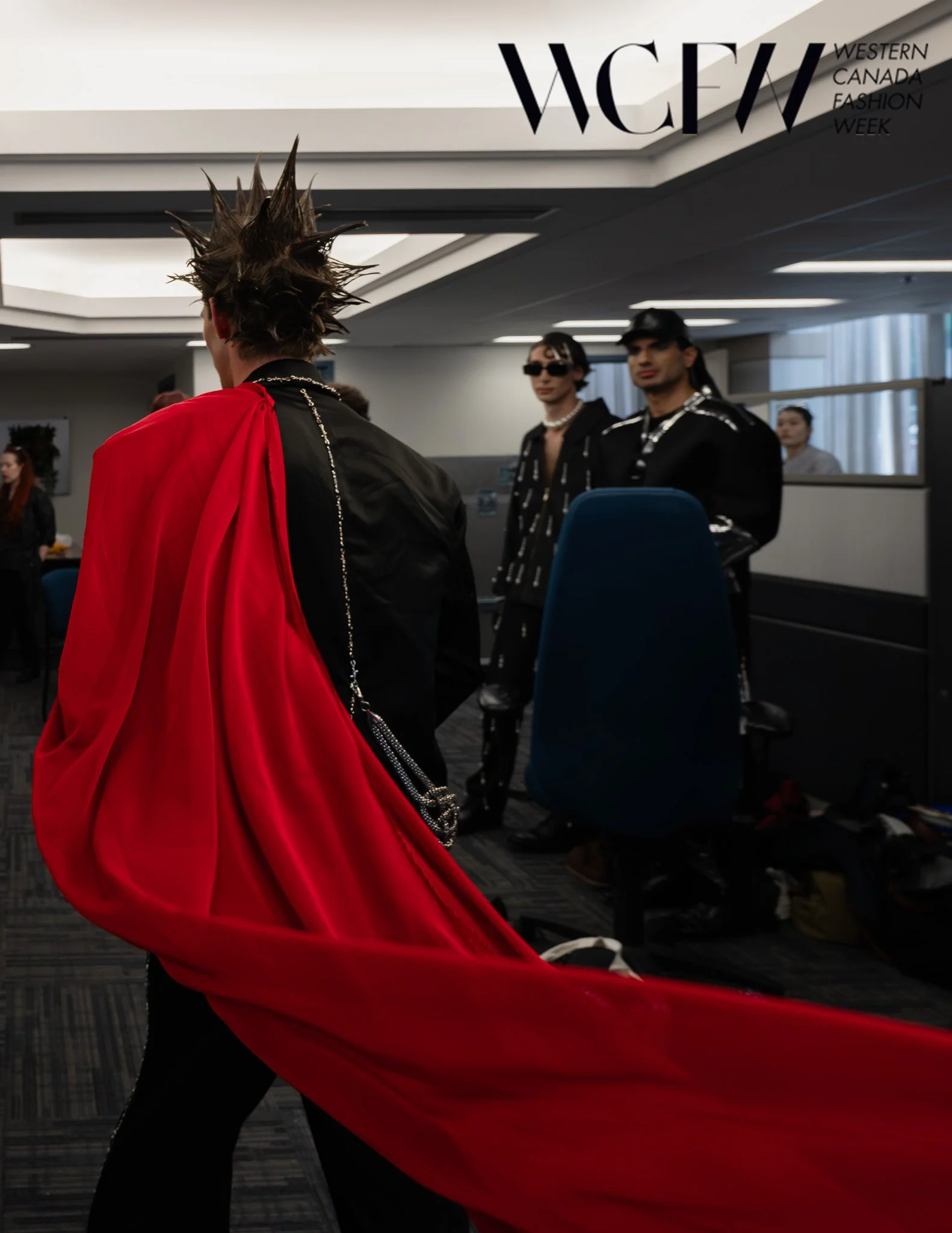 Person with spiked hair wearing a red cape at Western Canada Fashion Week, with two other models in the background.
