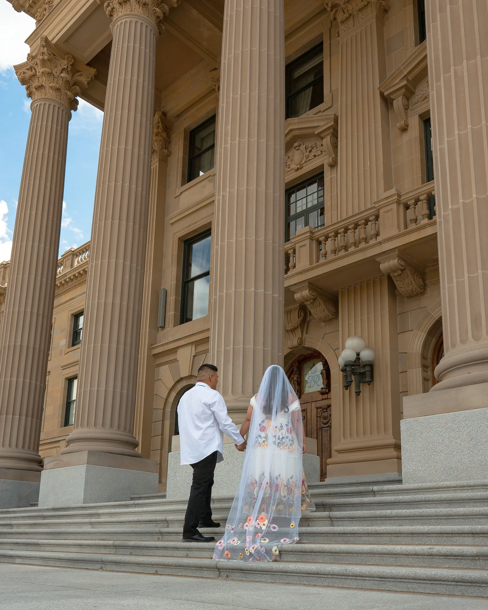 A bride and groom holding hands and standing on the steps of a grand, neoclassical building with large columns and ornate architecture.