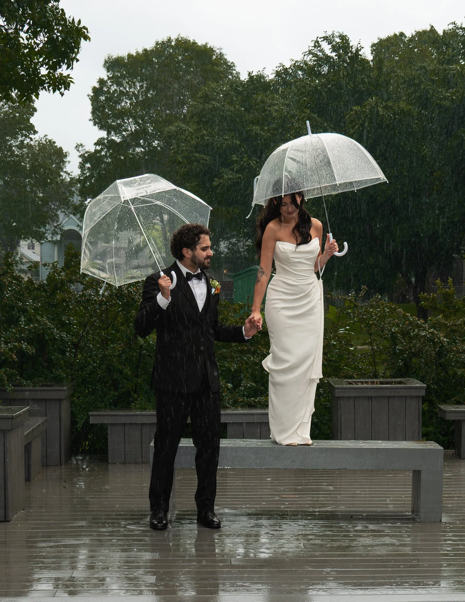 A couple dressed in formal wedding attire holding hands and standing on a platform outdoors in the rain, each holding a clear umbrella. The woman wears a white strapless dress, and the man wears a black tuxedo. They are surrounded by trees and a clou