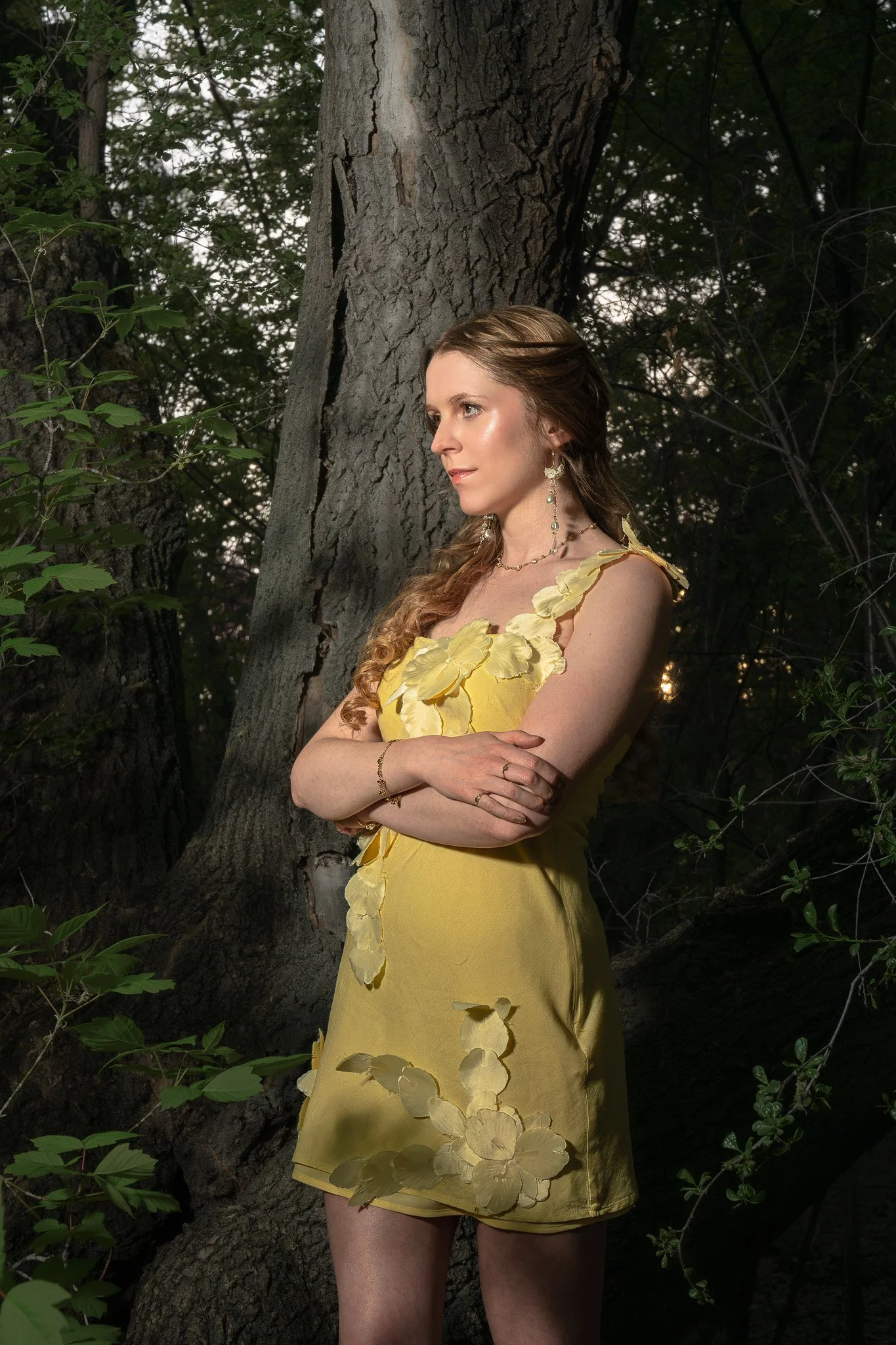 A woman in a yellow dress with floral details, earrings, and necklace, standing outdoors by a tree with dark green foliage.
