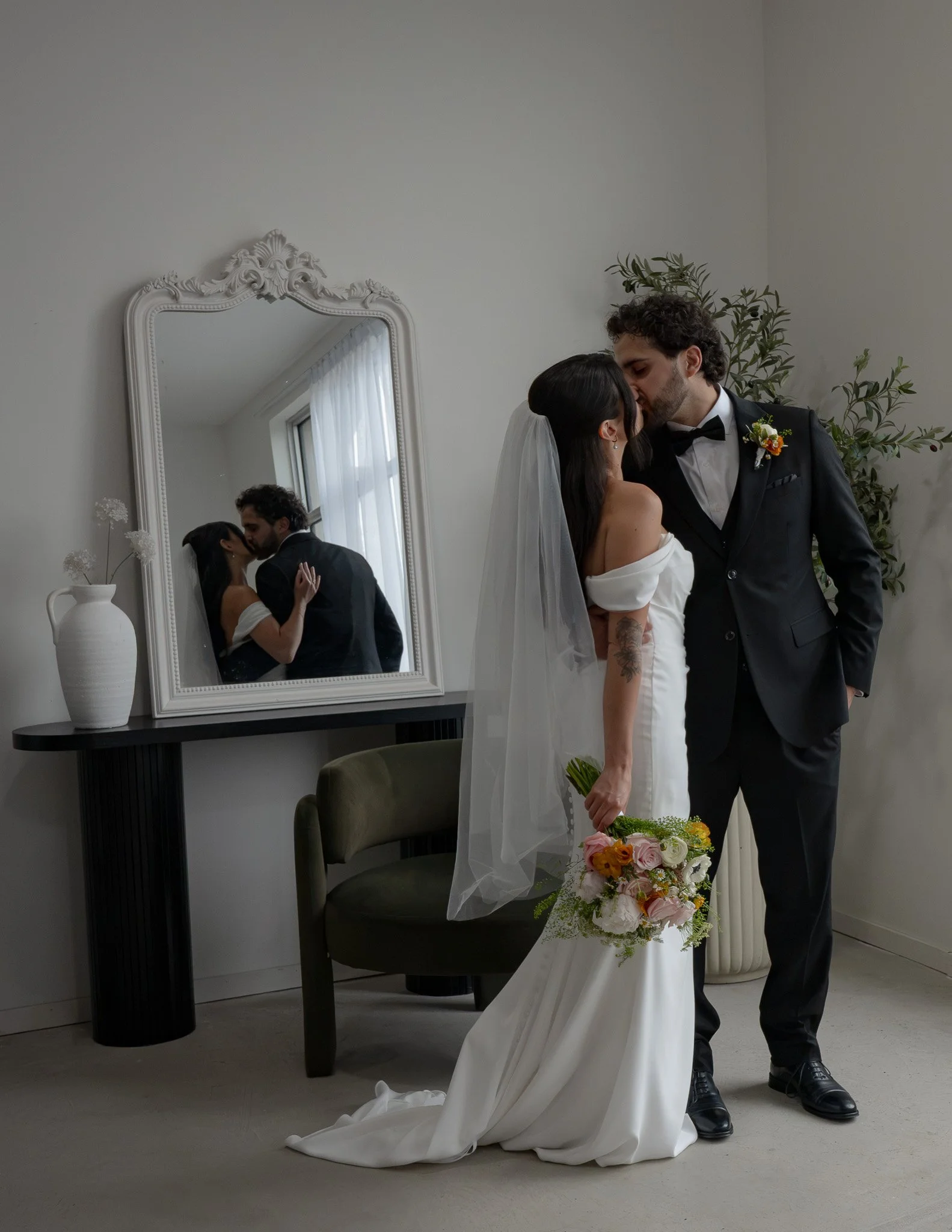A bride and groom are sharing a kiss in a mirror reflection, with the bride holding a bouquet of flowers and wearing a white dress & veil, and the groom in a black tuxedo with a flower boutonniere, in a minimalistic indoor setting.
