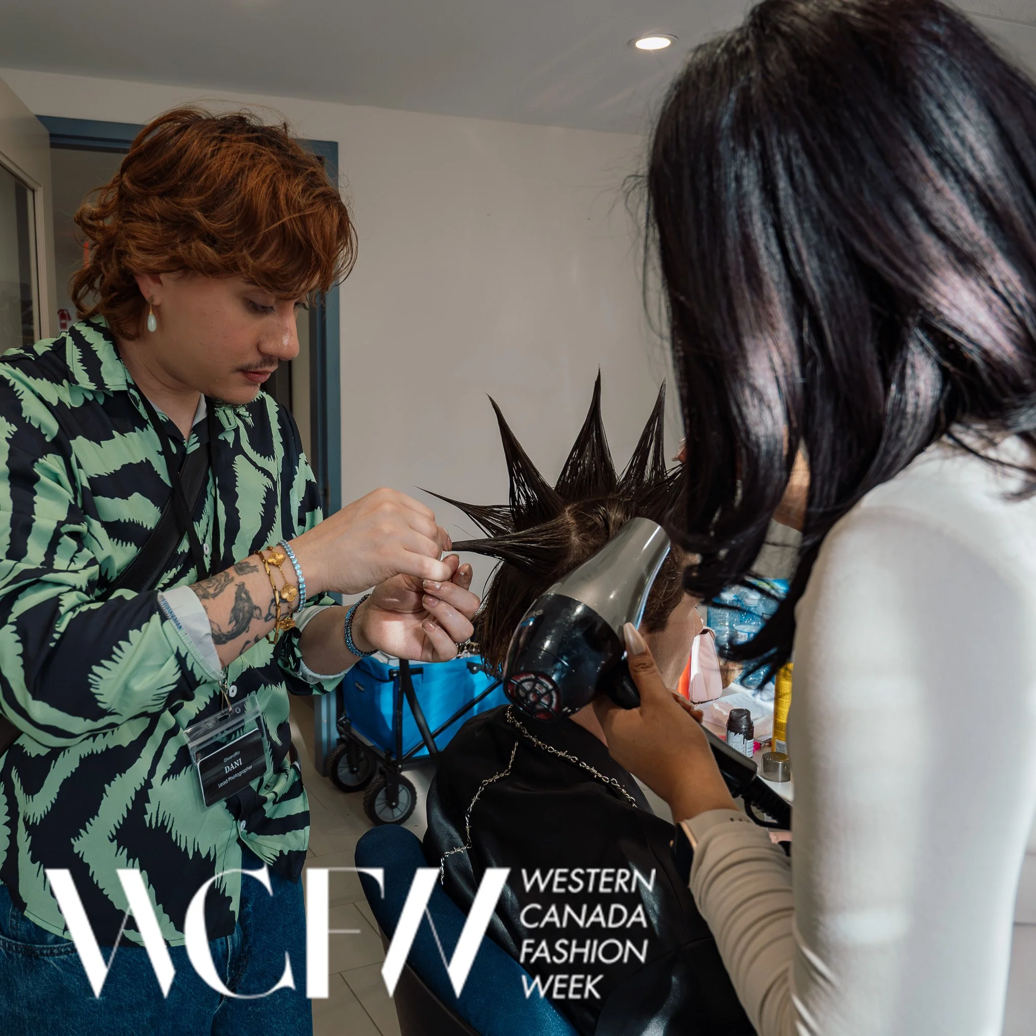 A hairstylist with tattoos and wearing earrings is sectioning and styling a woman's hair with foil during a hair treatment at a salon. The woman is seated with a black top and a chain strap.