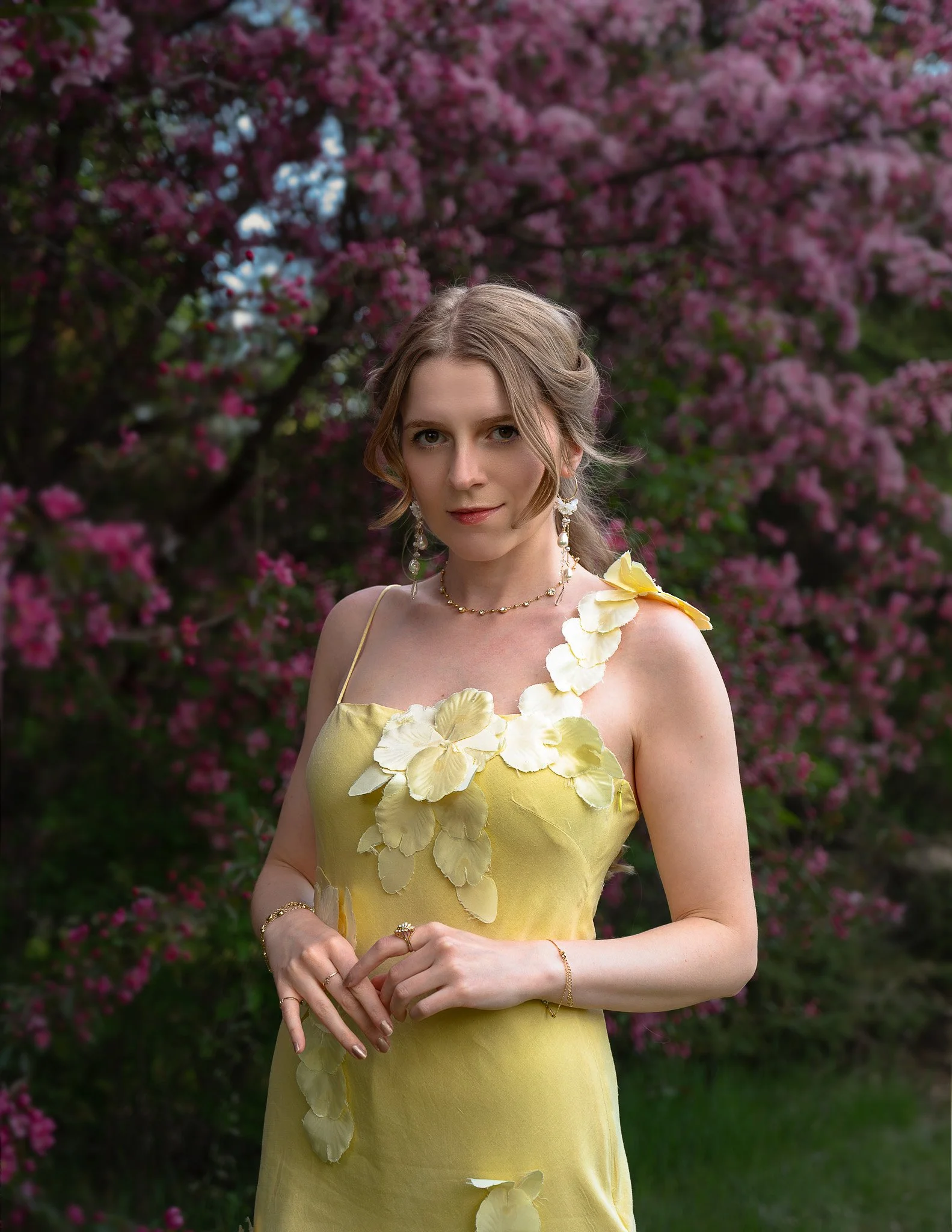 A young woman in a yellow dress with petal details standing outdoors in front of blooming pink flowers.