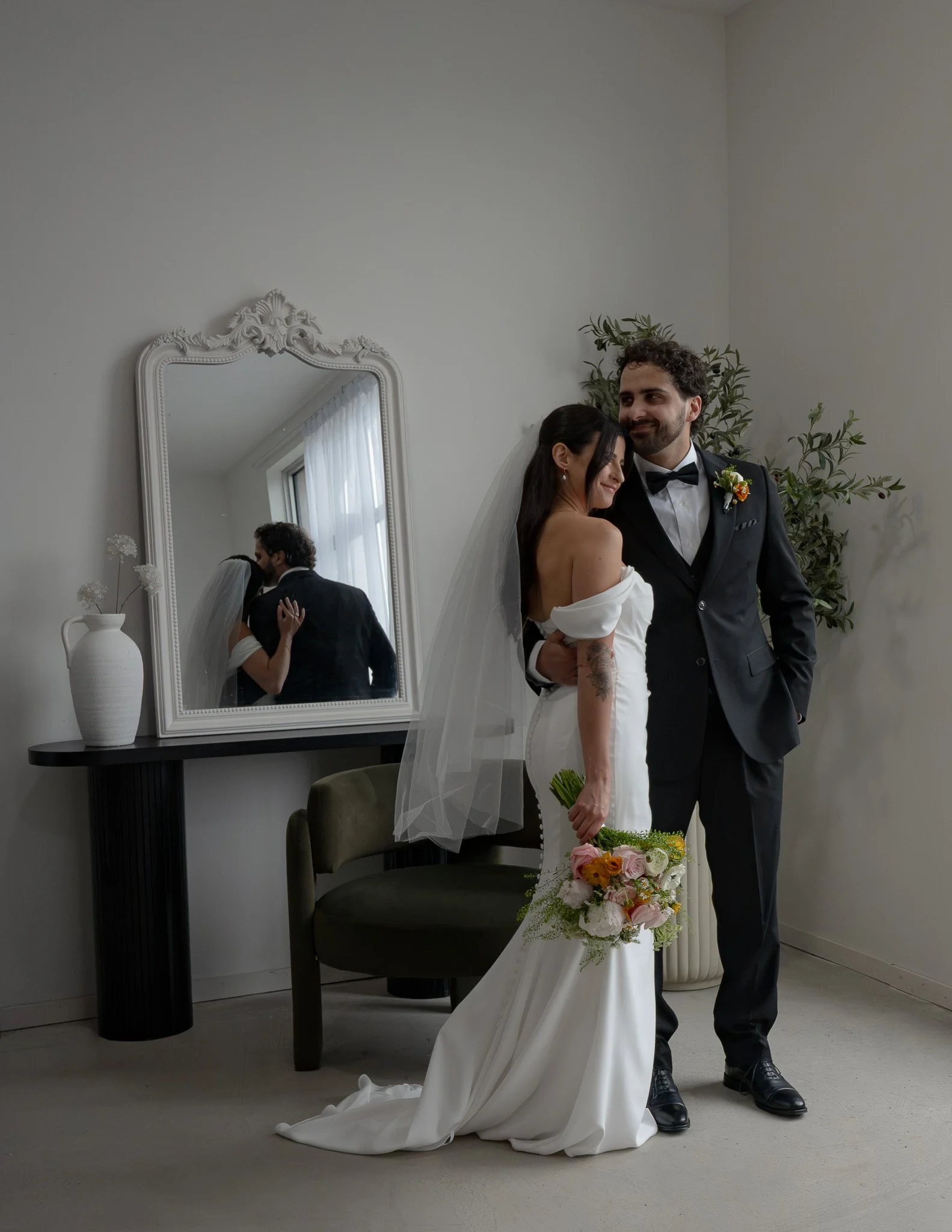 A bride and groom embracing near a mirror in a room with minimal decor. The bride holds a bouquet of flowers. The mirror reflects their image, and there is a vase with flowers on a table.