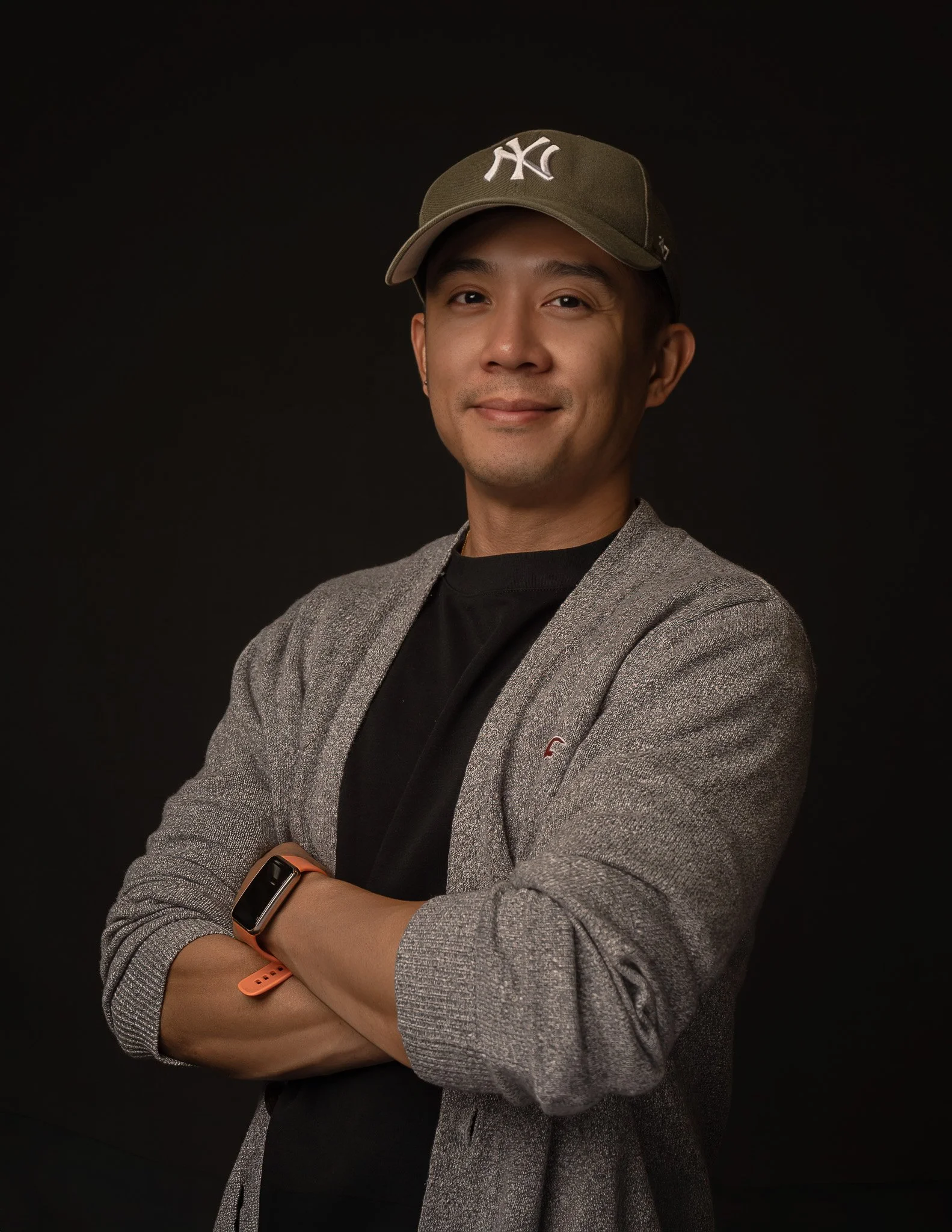 A young man with a slight smile wearing a gray blazer, a black shirt, a New York Yankees cap, and a smartwatch, standing against a black background.