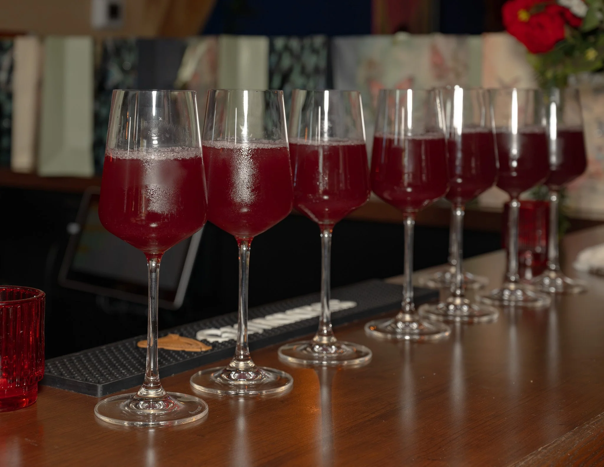A row of seven wine glasses filled with red wine on a wooden table, with a blurred bookshelf and flowers in the background.