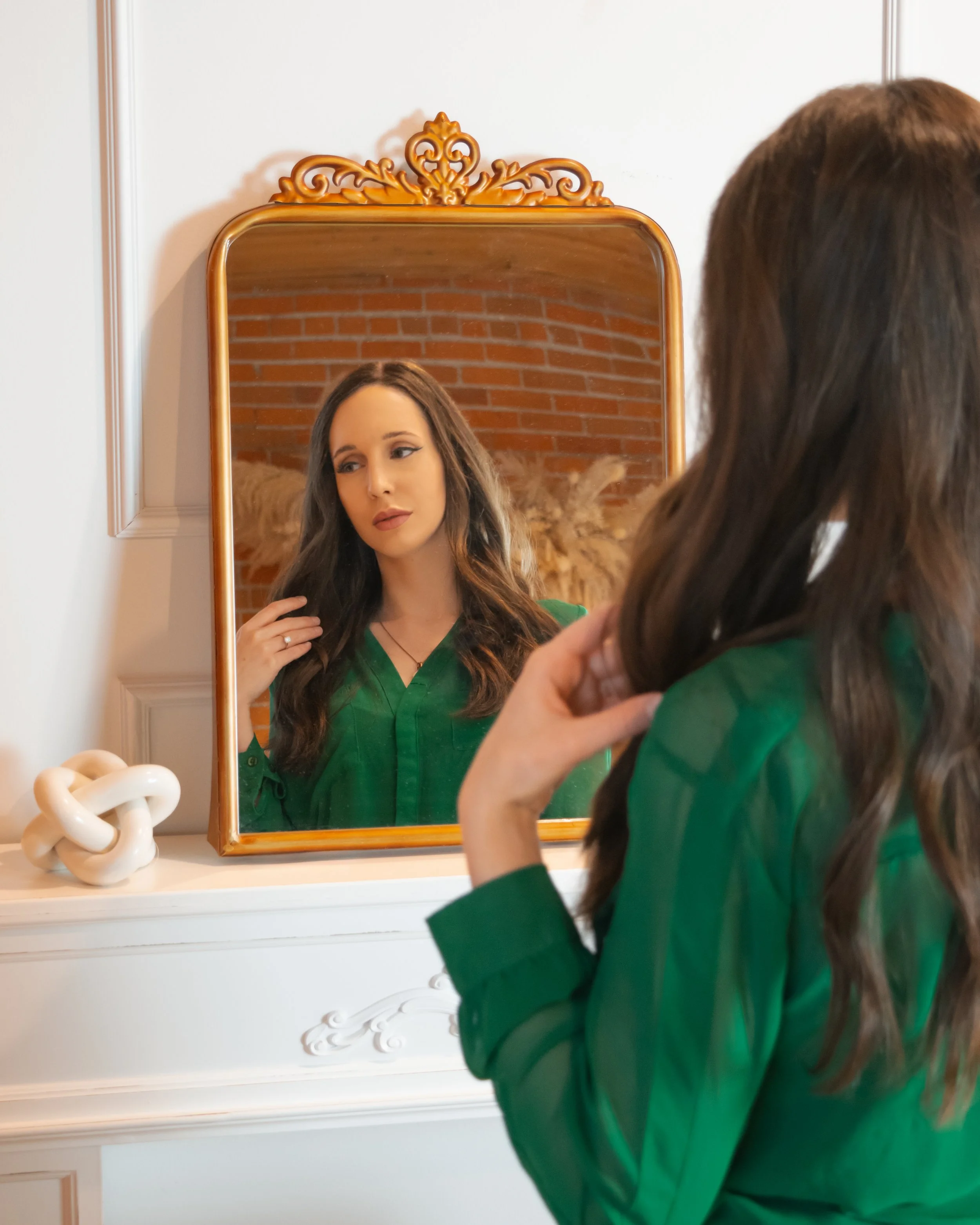 A woman with long brown hair looking at her reflection in a vintage mirror with a wooden frame, wearing a green blouse, in a room with white walls and a brick wall visible in the mirror's background.