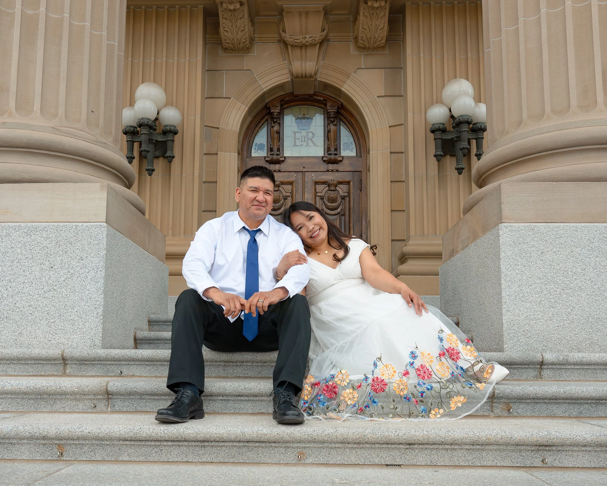 A couple sitting on the steps outside a large, ornate building with columns and decorative stonework. The man is wearing a white shirt and a blue tie, and the woman is wearing a white dress with a floral skirt. They are smiling and holding hands.