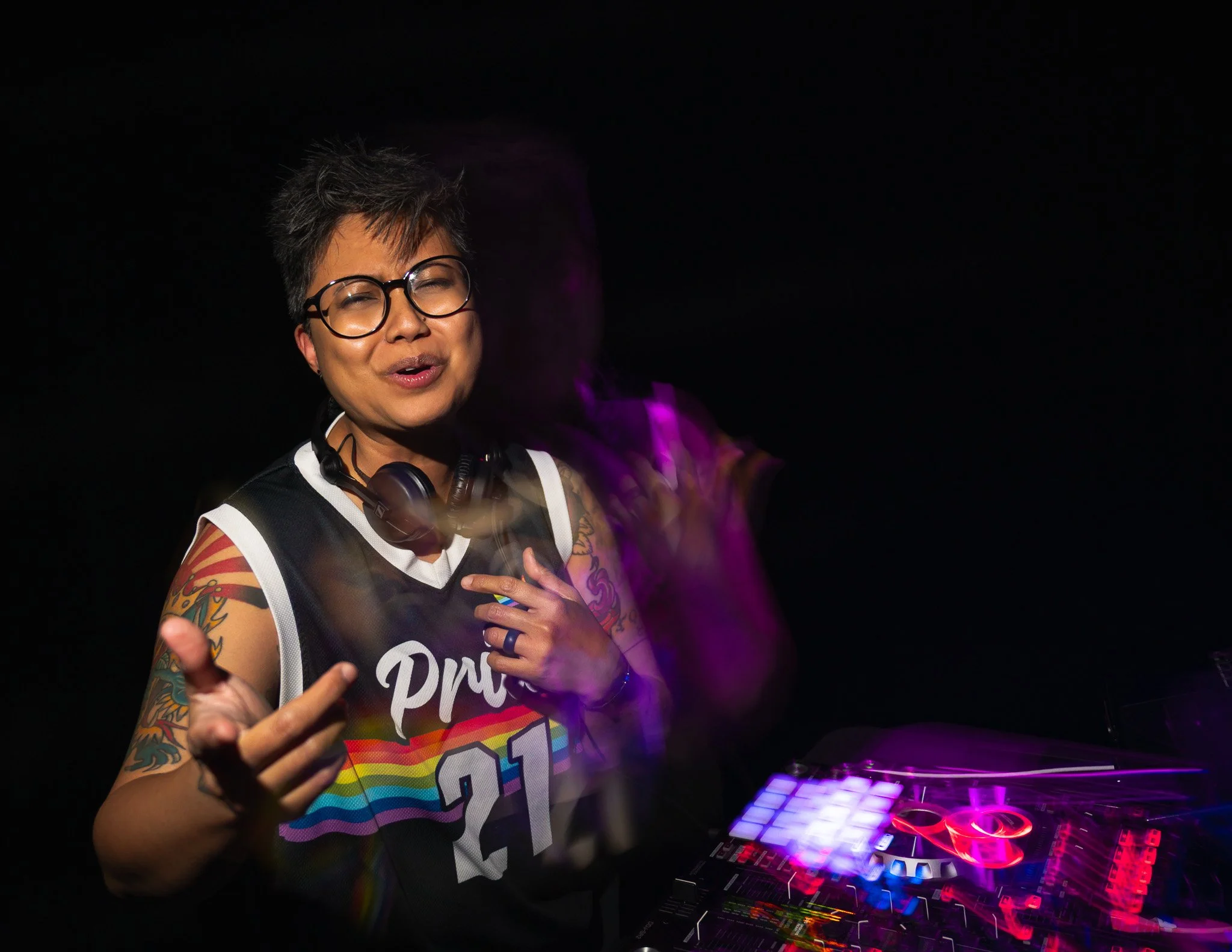 A person with short gray hair, glasses, and colorful tattoos on their arms, wearing a rainbow-colored party shirt, appears to be DJing or performing with a DJ controller and colorful LED lights in a dark setting.