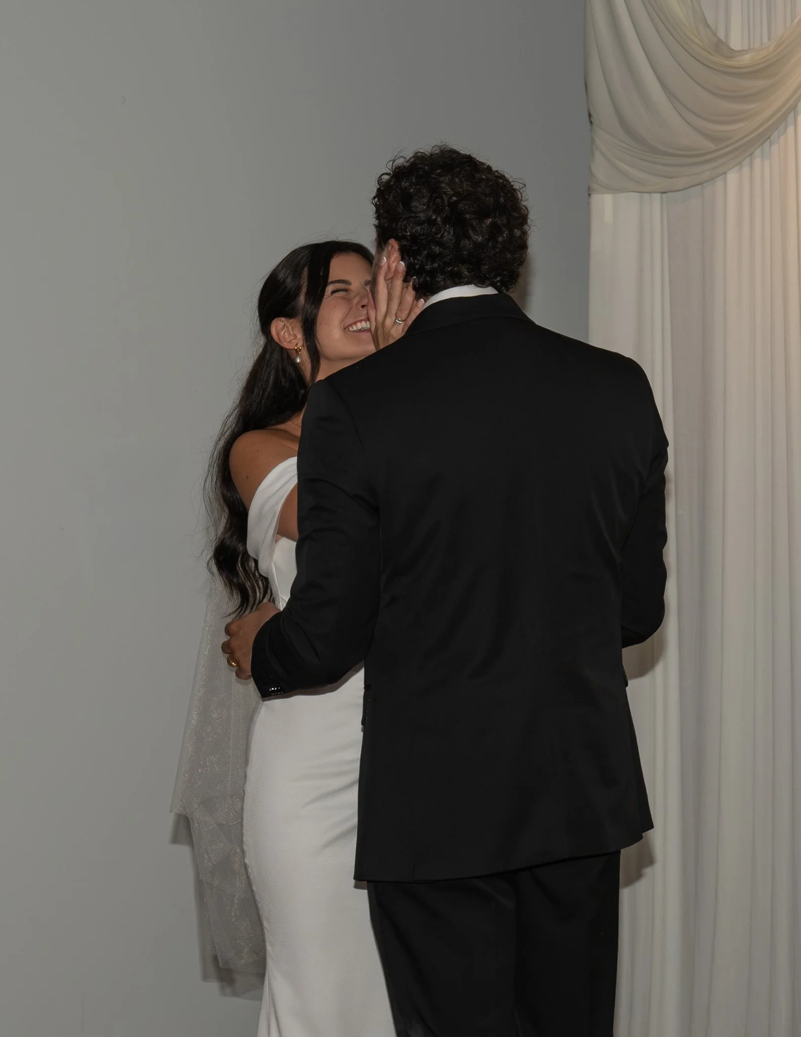A bride and groom share a kiss during their wedding ceremony, with the bride smiling and holding the groom's face, and the groom wearing a black tuxedo.