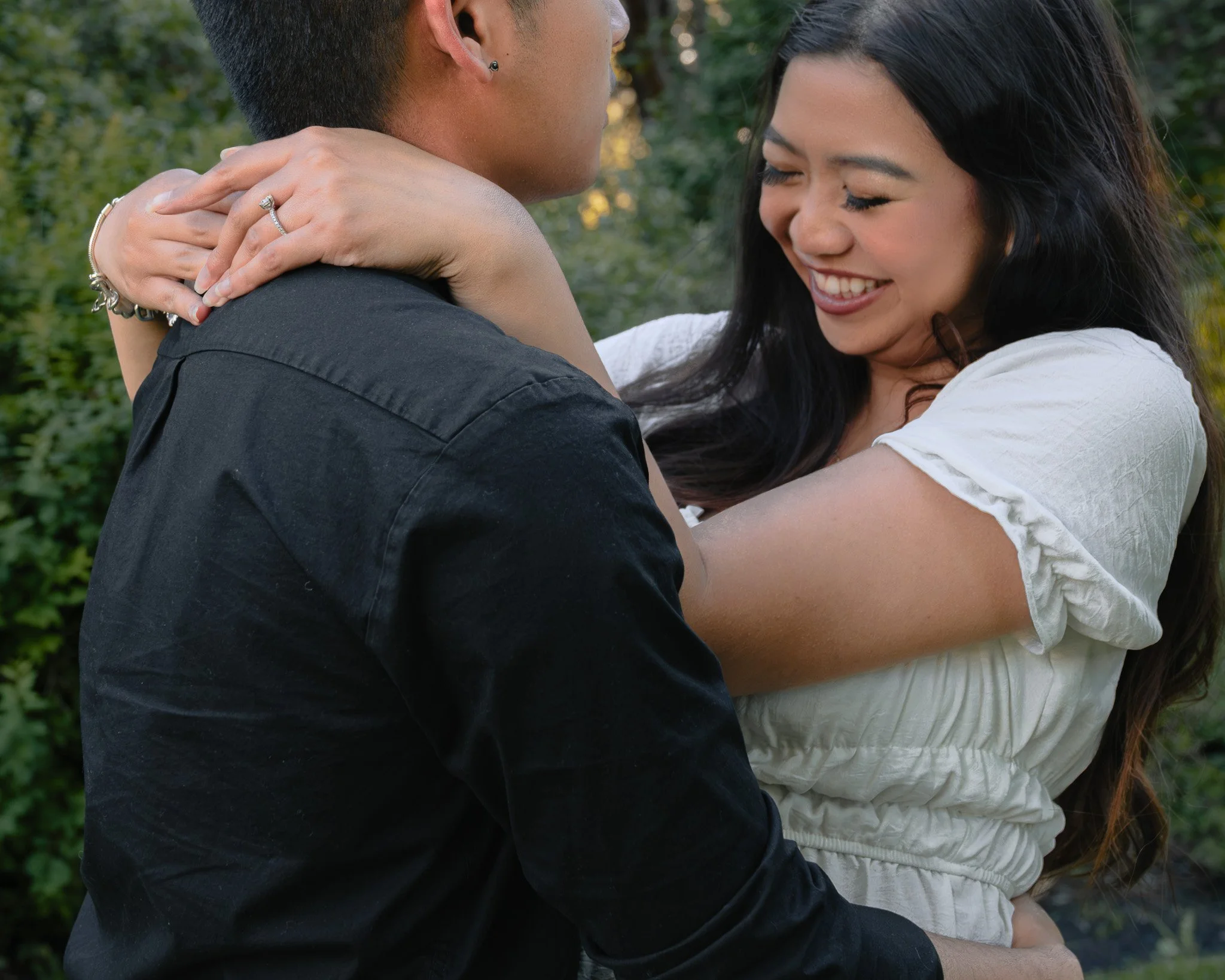 A young couple hugging outdoors, smiling and showing affection. The woman has long dark hair, and the man has short dark hair, wearing a black shirt. The woman has jewelry, including a ring and bracelets. The background is green foliage.