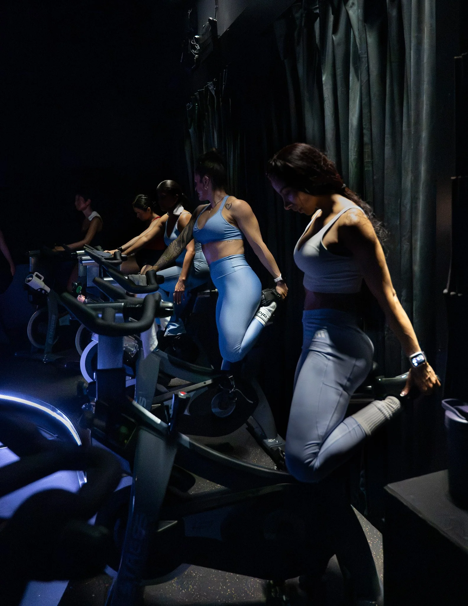 A group of women dressed in athletic wear exercising on stationary bikes in a dark gym with black curtains.