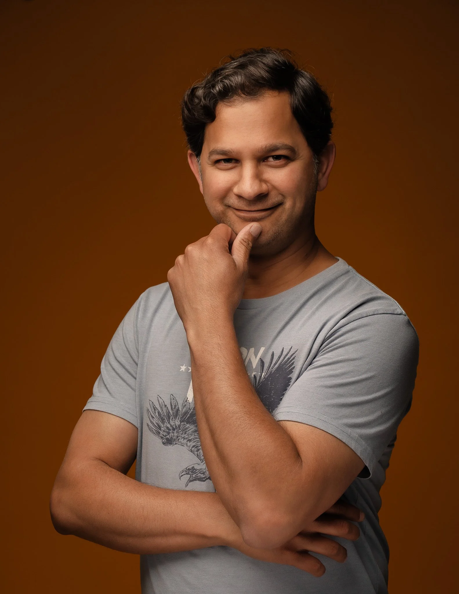 A man with dark wavy hair smiling, touching his chin with his right hand, crossed arms, wearing a gray tribal eagle t-shirt, against a brown background.