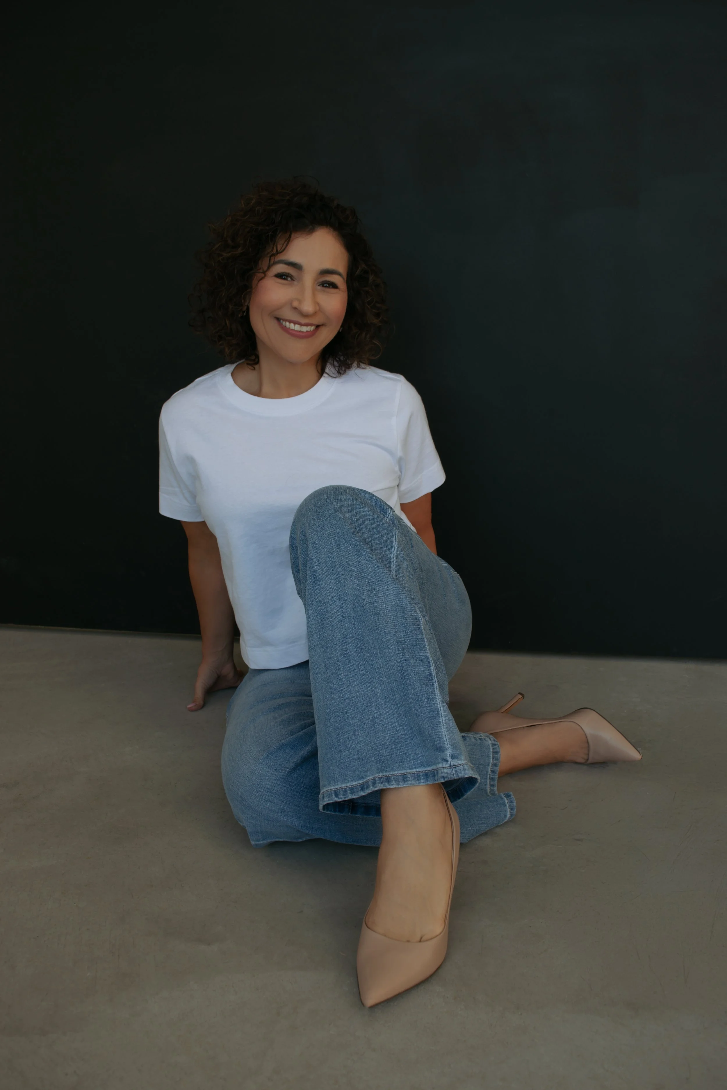 Woman with curly hair smiling while sitting on the floor against a black wall, wearing a white t-shirt, blue jeans, and nude heels.