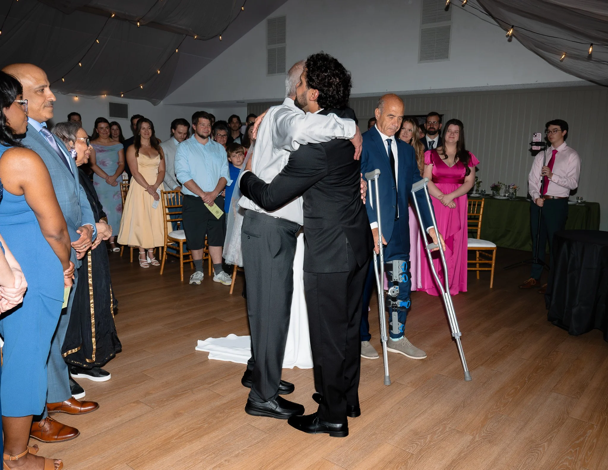 A group of people standing and watching a hug between two men, one in a suit with dental crutches, and the other in a black jacket, at an indoor event or celebration.