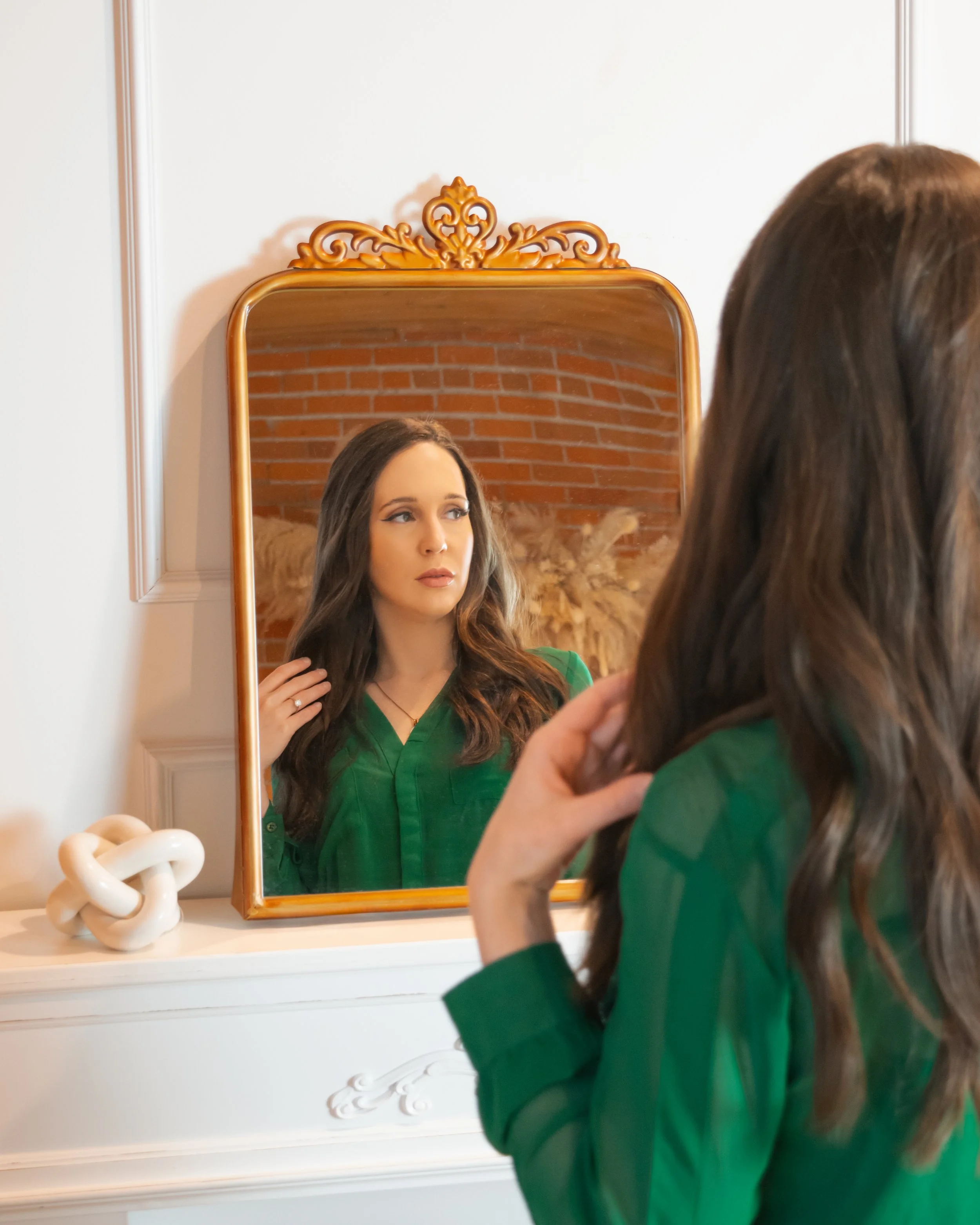 A woman with long brown hair in a green blouse looking at her reflection in a mirror with a decorative wooden frame, touching her hair with her right hand, against a brick wall background.