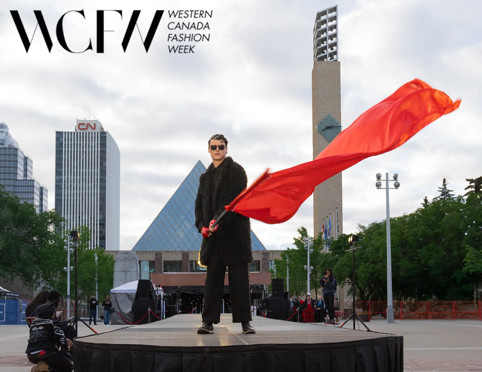A person holding a red cloth flag standing on a stage outdoors in front of a cityscape with tall buildings and cloudy sky, during Western Canada Fashion Week.