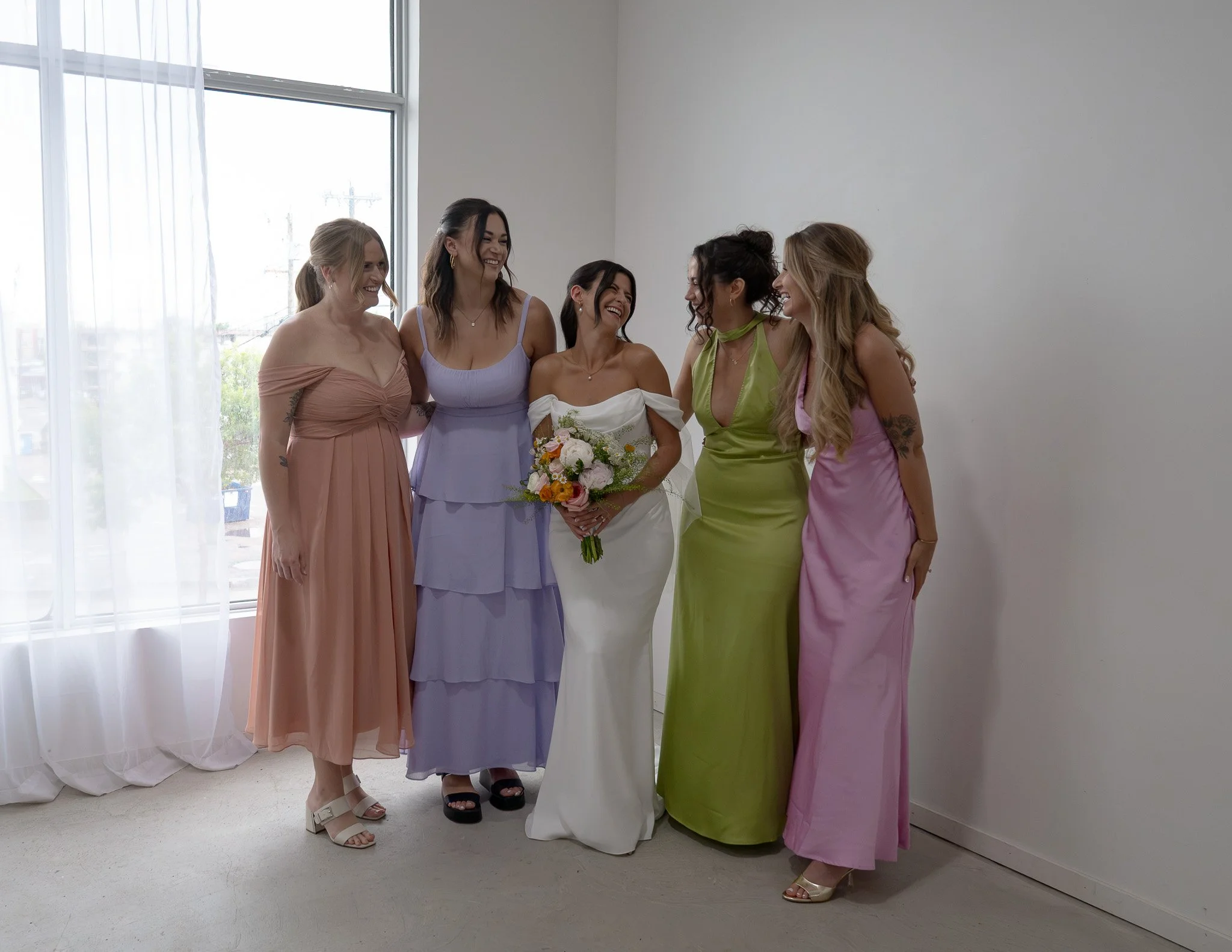 Five women dressed in colorful dresses smiling and posing for a photo indoors near a large window with sheer curtains.